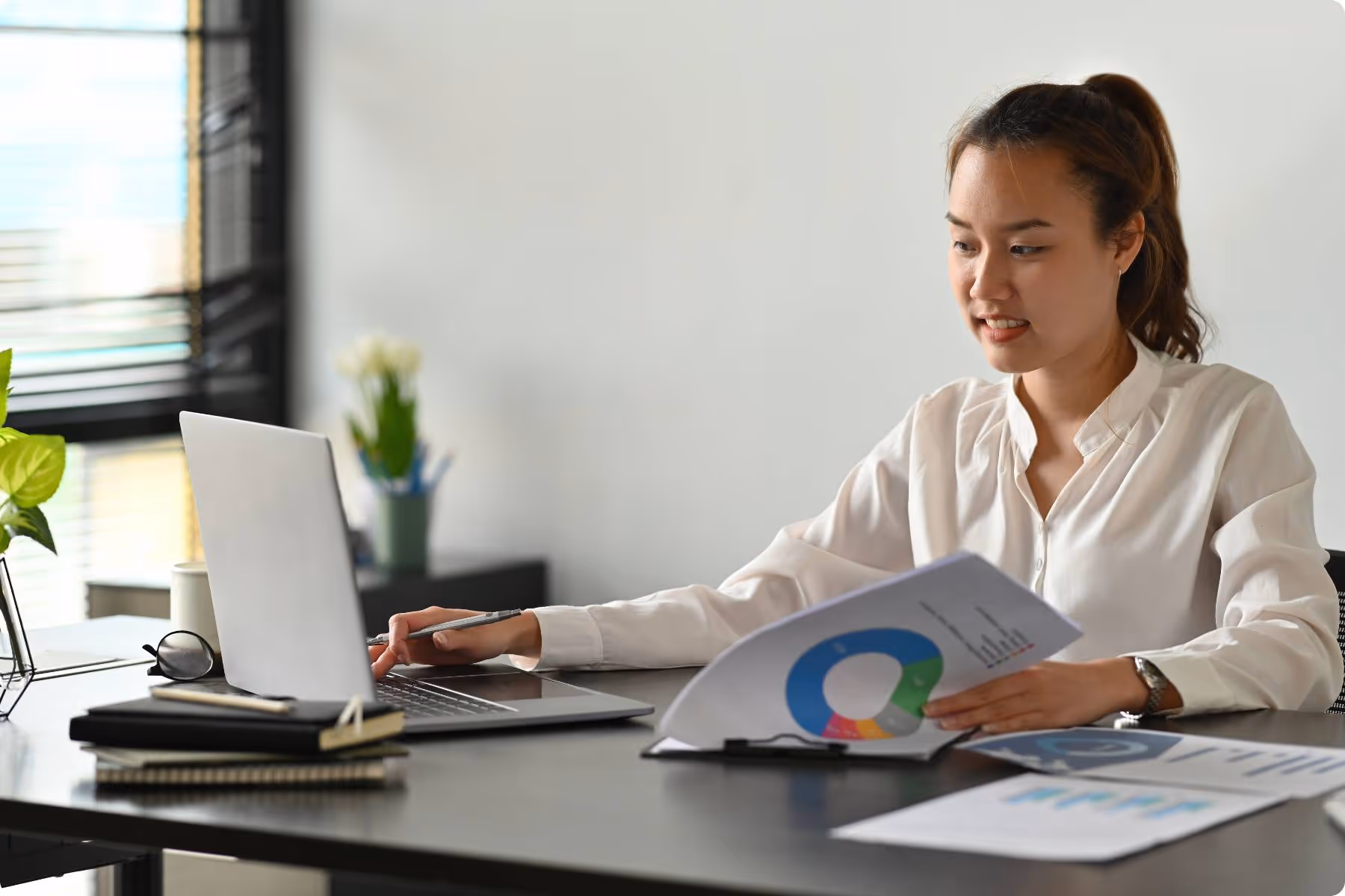 Eine Frau arbeitet an einem Schreibtisch mit Laptop und hält ein Diagramm in der Hand. Auf dem Tisch liegen weitere Dokumente und Notizbücher in einem modernen Büro.