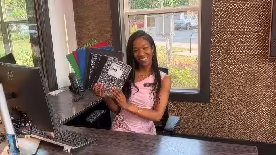 A woman holding folders and a composition notebook at a work station, smiling.