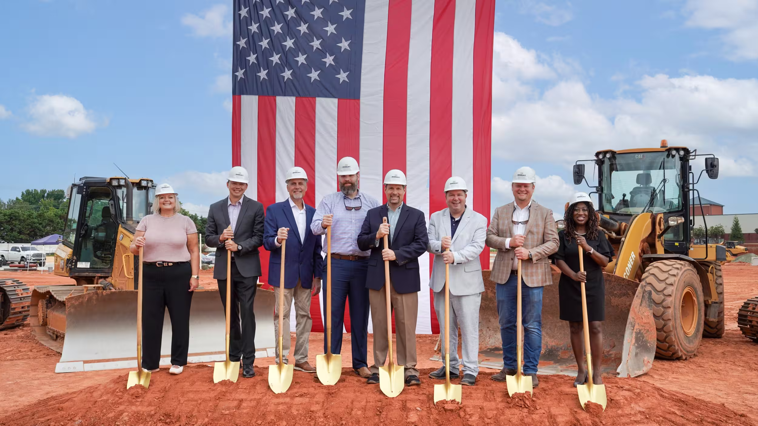 People standing in a line with shovels in front of them going into the dirt with an American Flag behind them.