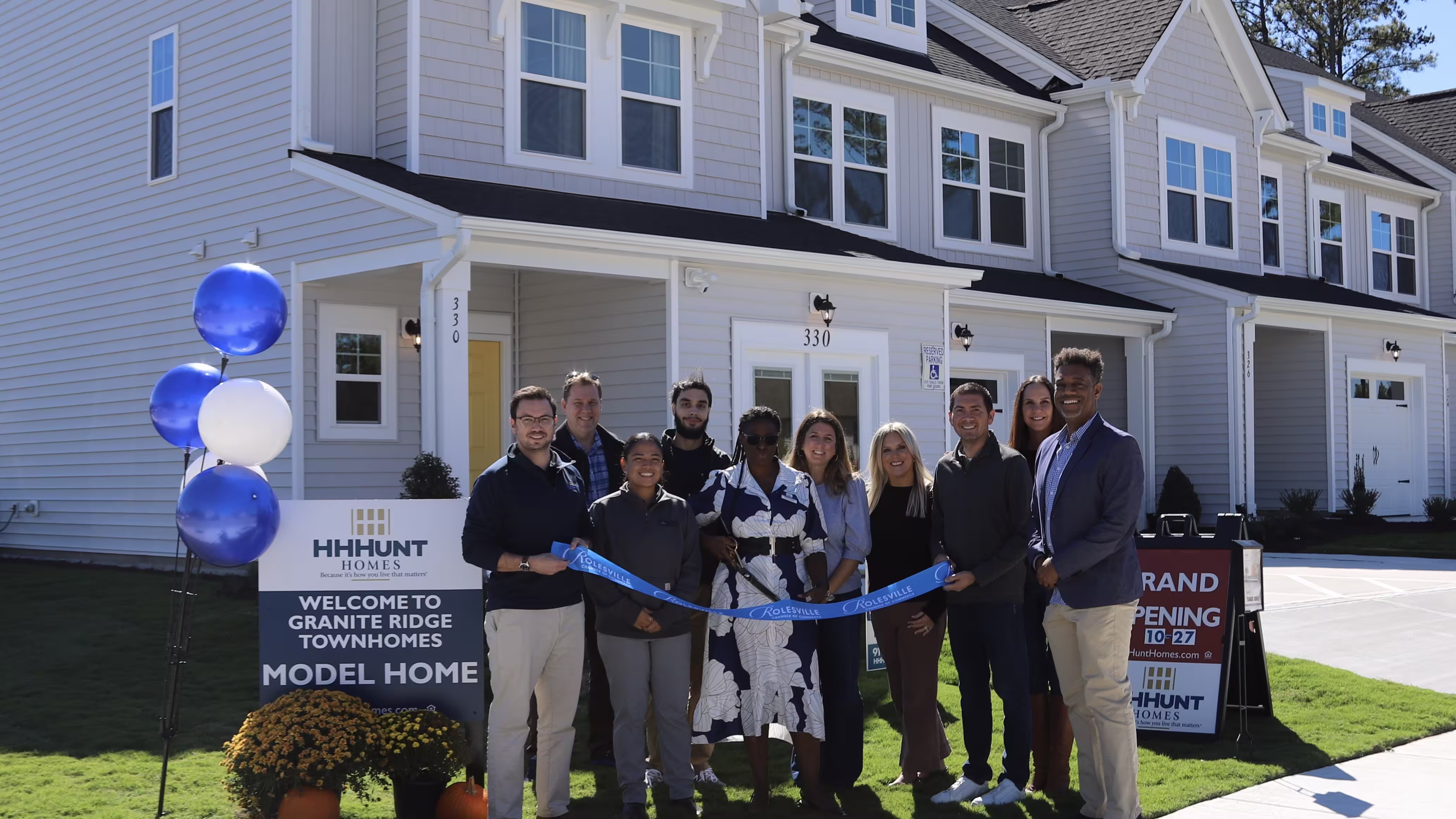 Group of people standing with a ribbon and balloons in front of a strip of townhomes.