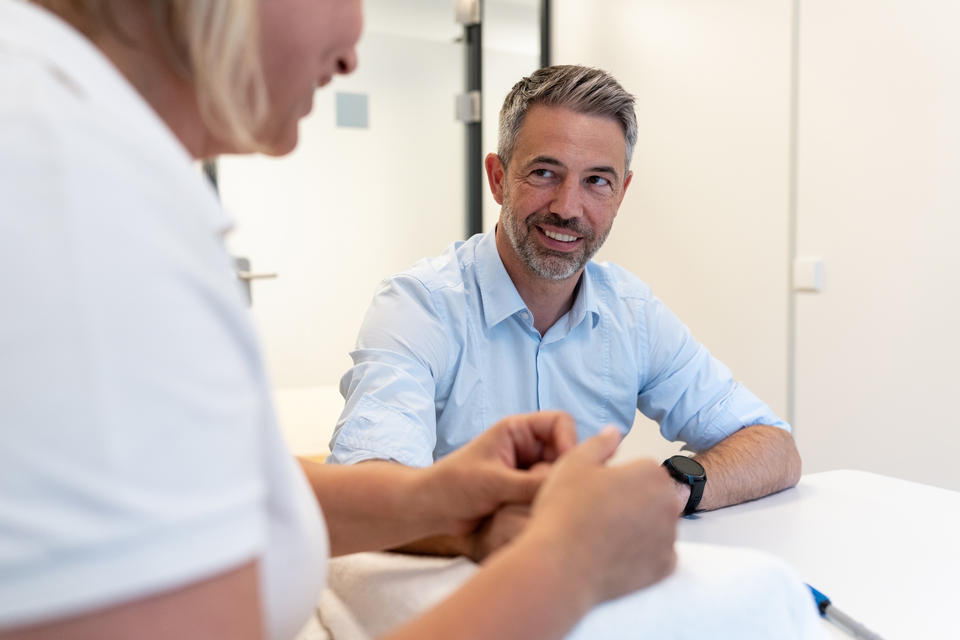 Therapeutin führt Handübungen mit einem jungen Mann am Tisch in einer Praxis durch, beide tragen Mundschutz.