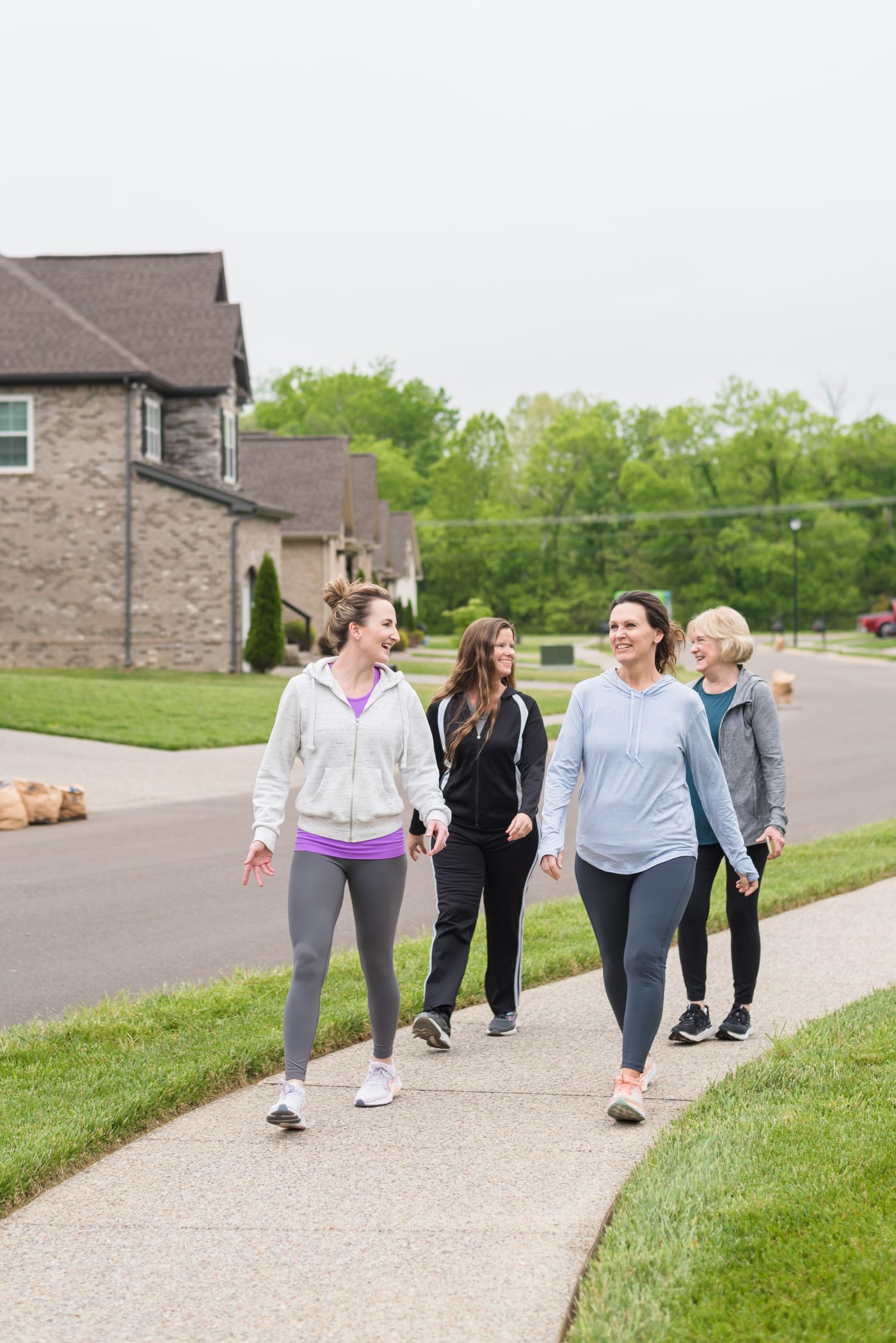 Four women of different ages walking together on a neighborhood sidewalk, talking and laughing.