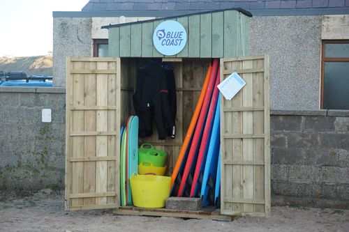 The Blue Coast Self-Service Surf Hire Kiosk at Cullen Beach with its doors open and a range of surfboards and wetsuits visible inside.