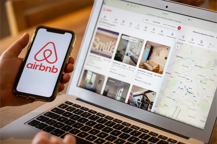 Hand holding a smartphone displaying the Airbnb logo, with the Airbnb rental listings website open on a laptop screen in the background, representing online property management services.