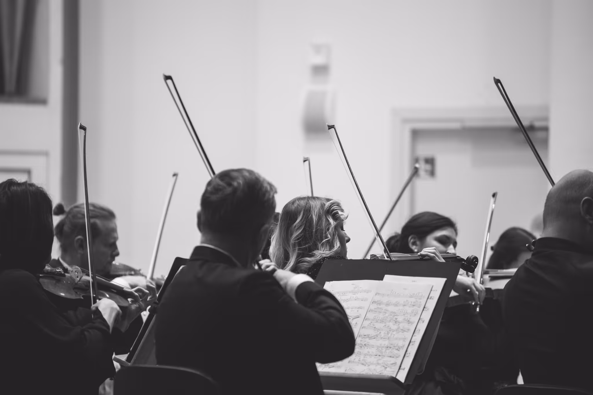 Black and white image of Sinfonietta Cracovia orchestra musicians playing violins, viewed from behind with sheet music in foreground.