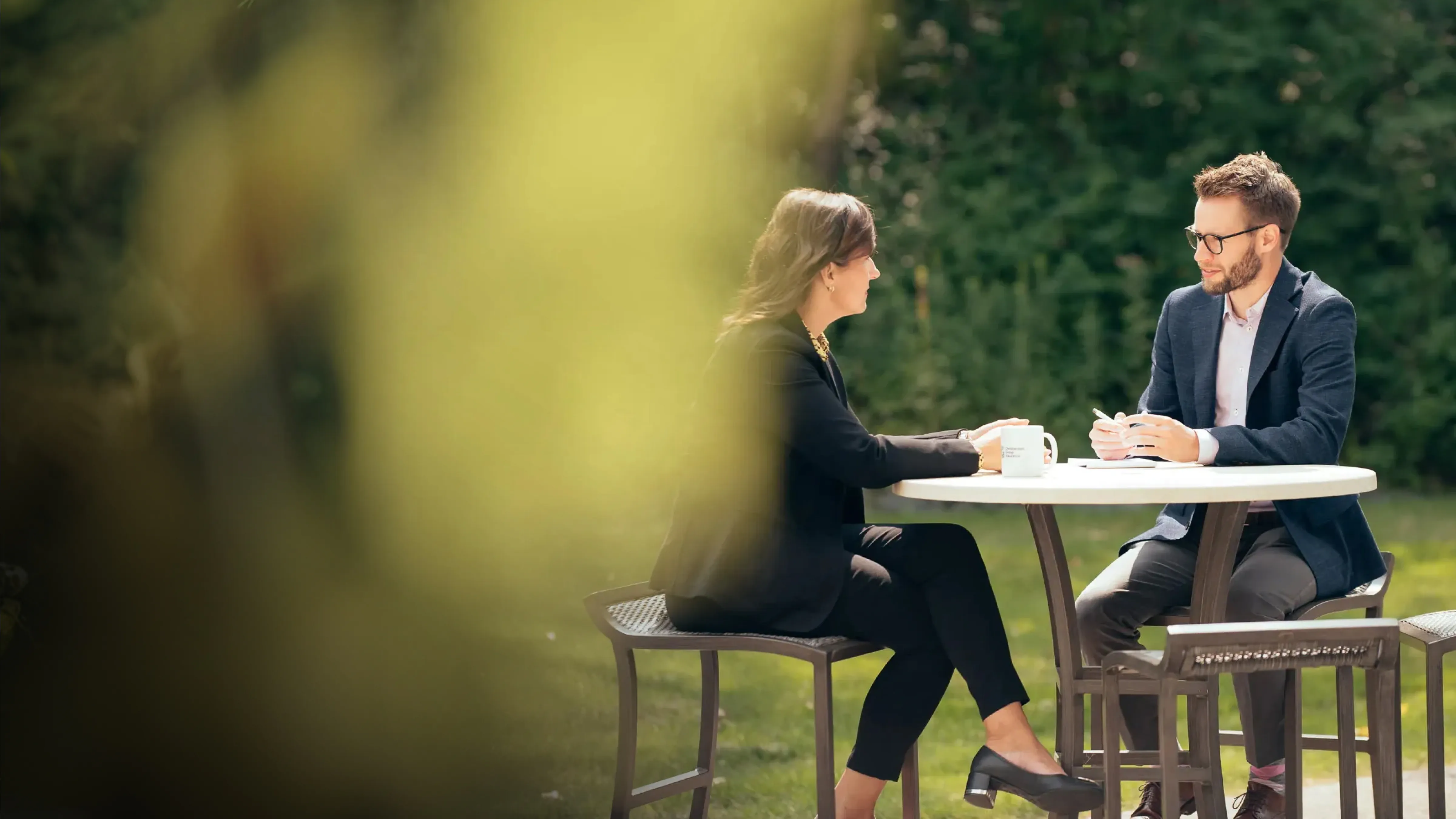 Two people sitting at an outdoor table having a conversation, holding coffee mugs.