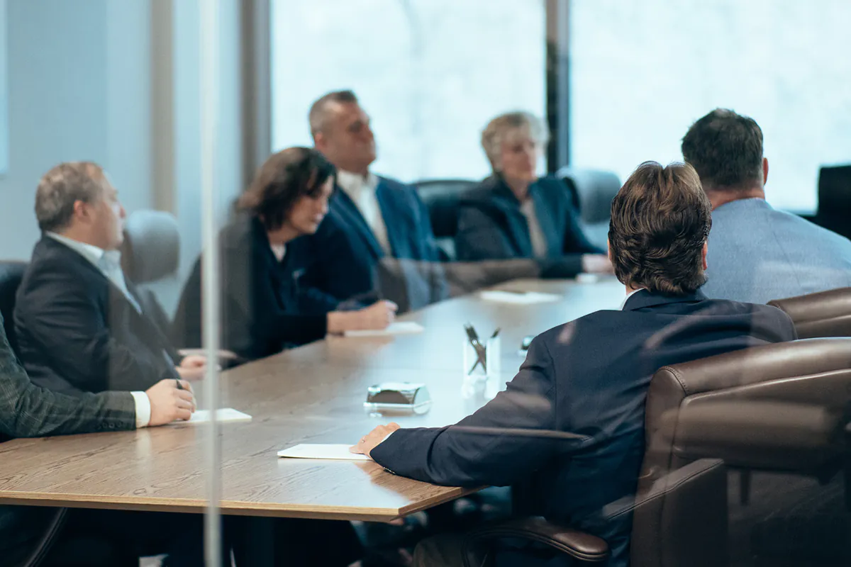 Group of business professionals seated around a large conference table in a meeting room, some taking notes, seen through a glass wall.