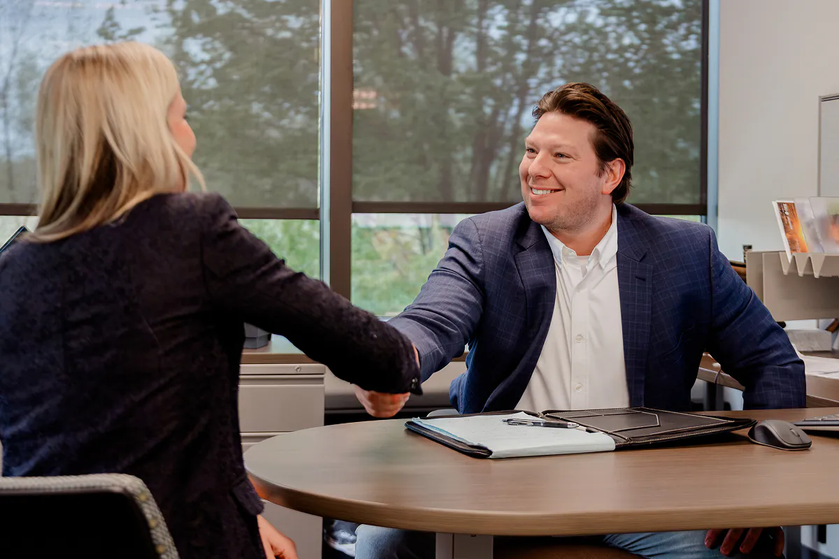 Two people shaking hands across a round office table, a smiling man in a blue blazer and white shirt faces a woman with blonde hair wearing a dark jacket, with a window showing trees in the background.