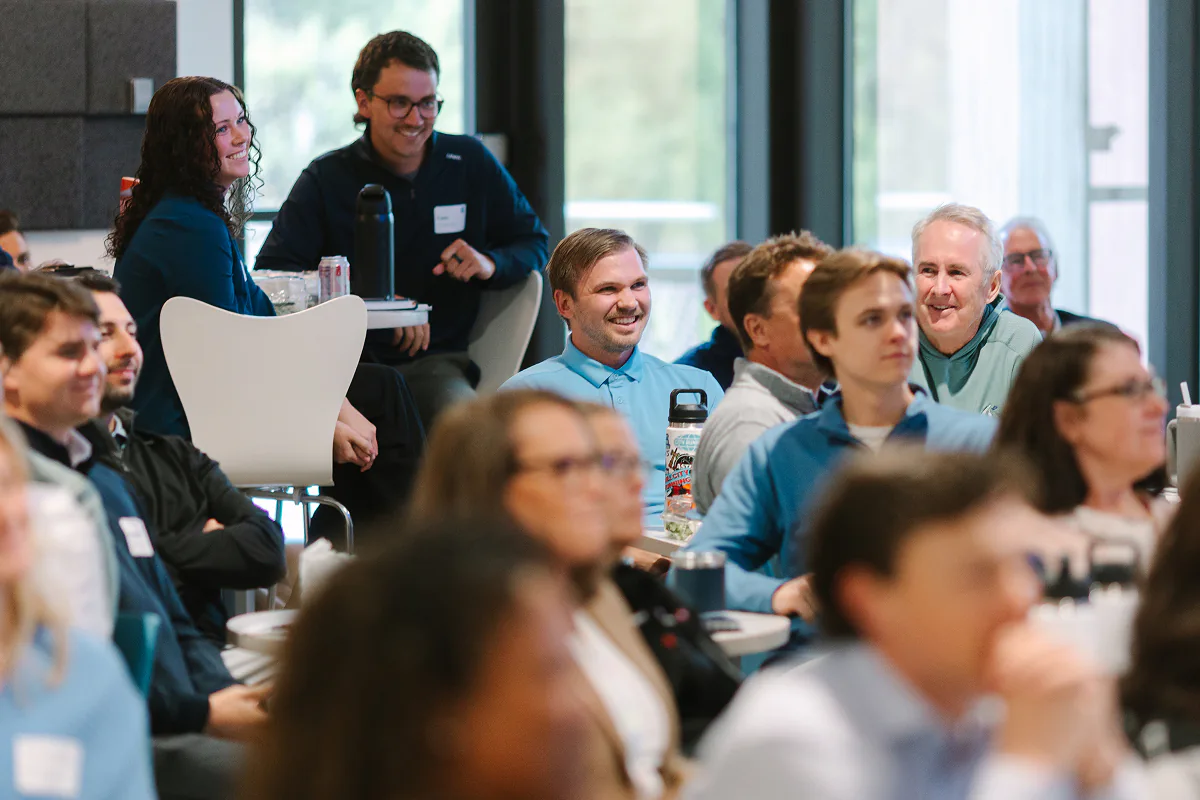 A diverse group of people seated indoors attentively listening and smiling during a meeting or presentation in a bright room with large windows.