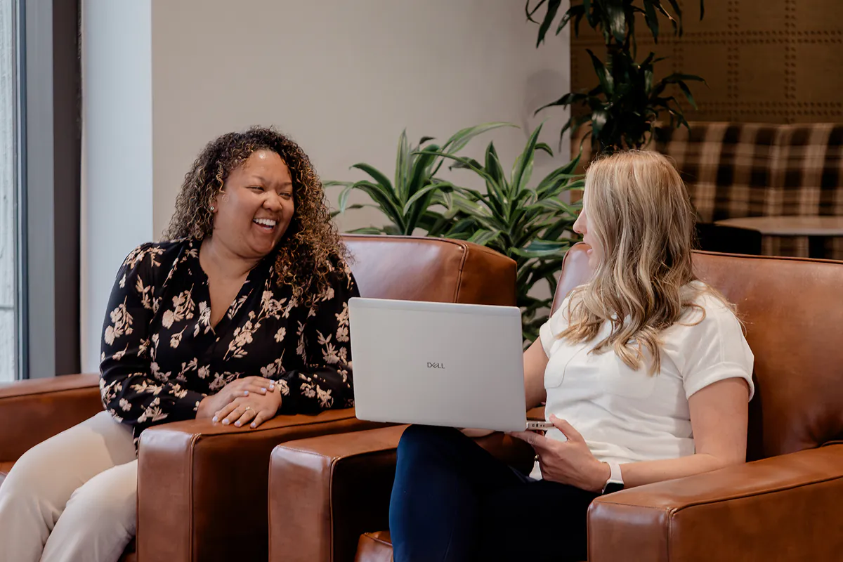 Two women sitting on brown leather chairs in an office setting, one holding a laptop and both smiling and engaging in conversation.