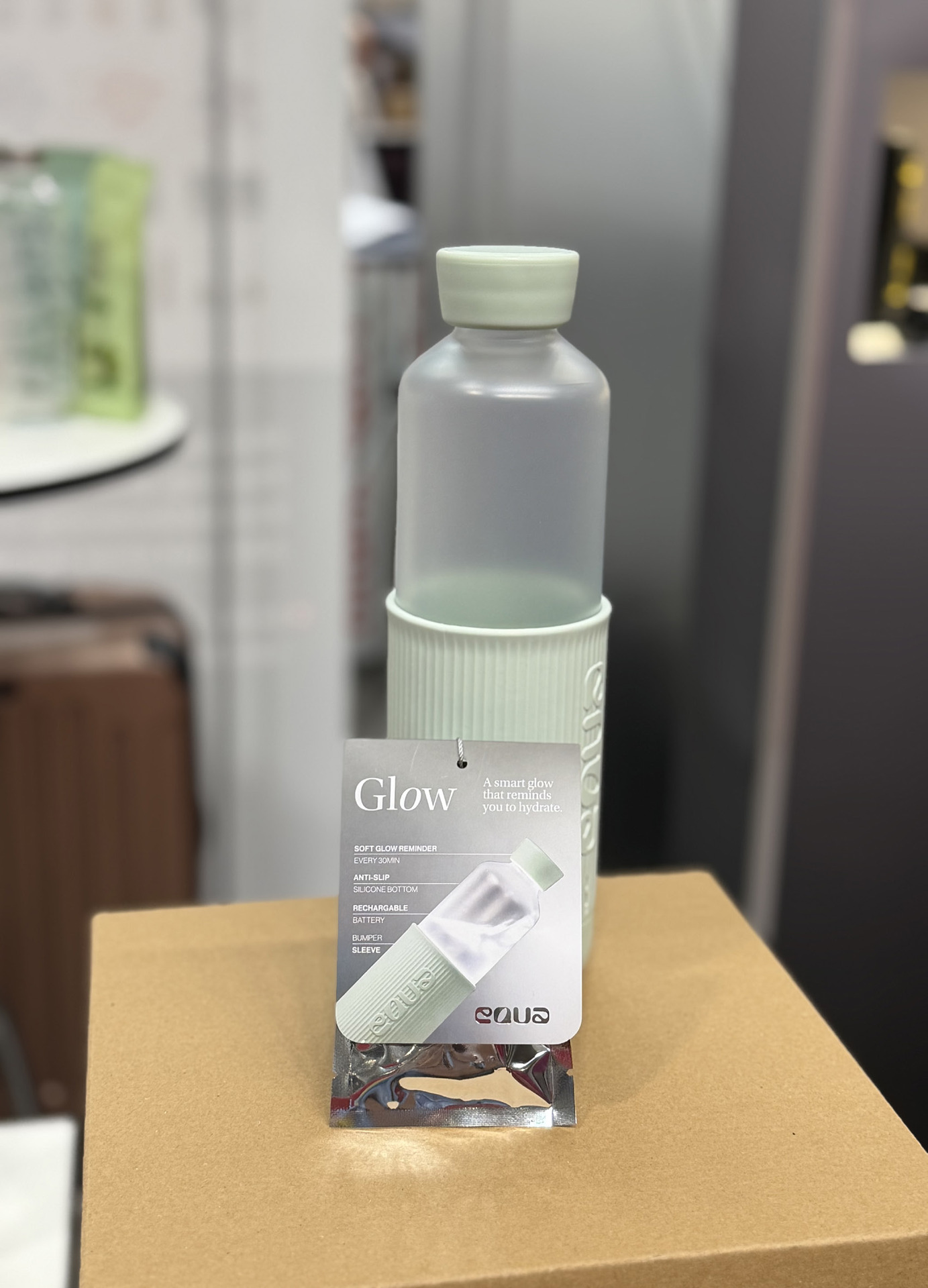 Glasflasche Glow Bottle mit mintgrüner Silikonhülle und Deckel auf einem Kartonsockel, davor die Produktverpackung