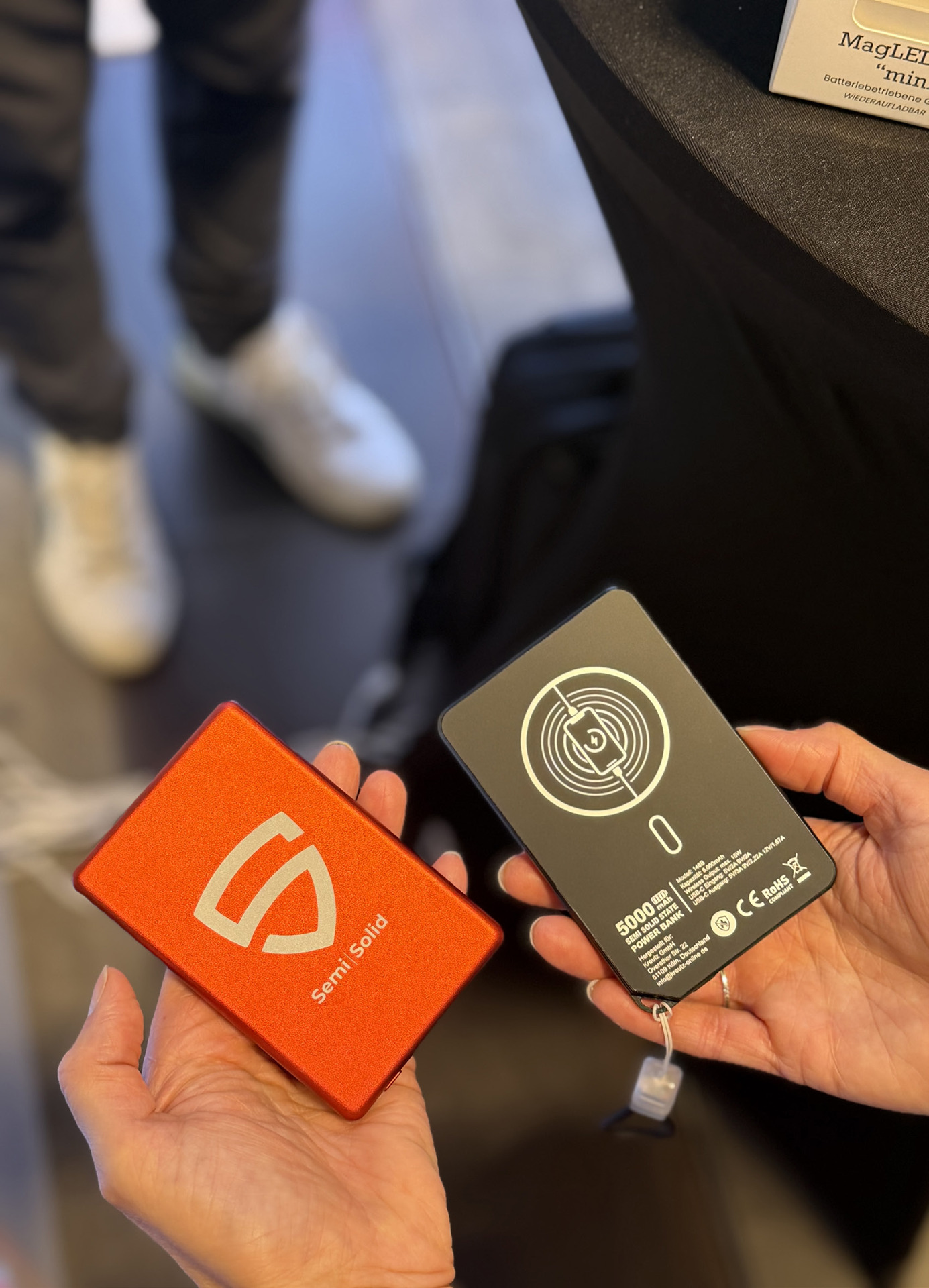 Zwei flache Powerbanks MagPower Safety in Orange und Schwarz, in den Händen von Messebesuchern