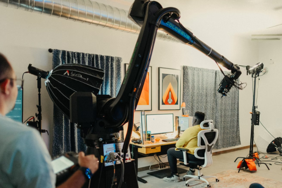 A robotic camera arm films a person at a desk in a modern studio setup; lighting equipment and crew members are present, capturing behind-the-scenes of a creative shoot.