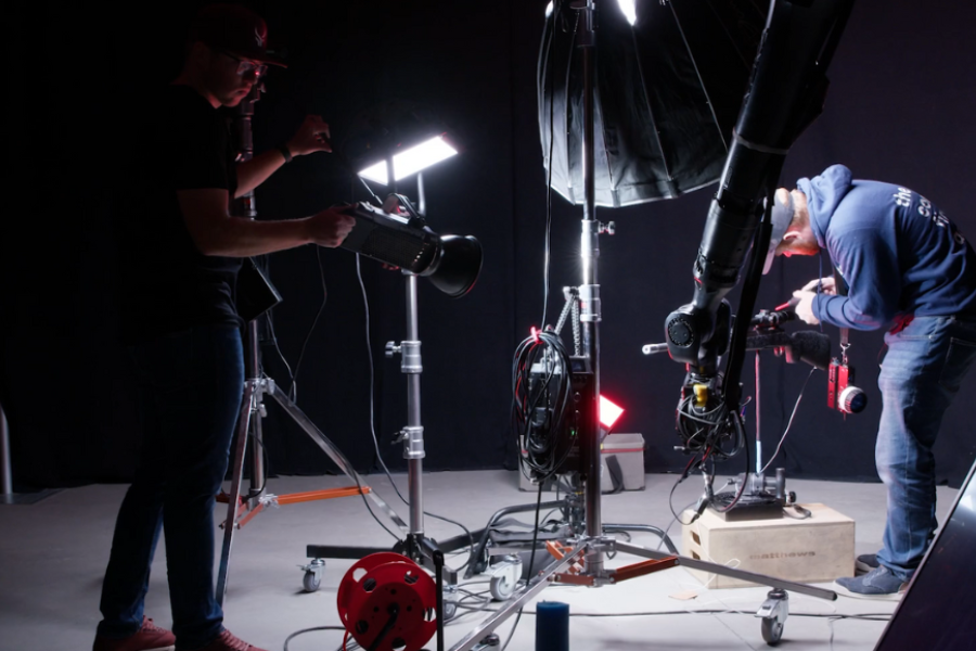 Two crew members working in a dark studio with professional lighting and a robotic camera arm; one adjusts a light while the other fine-tunes camera settings.