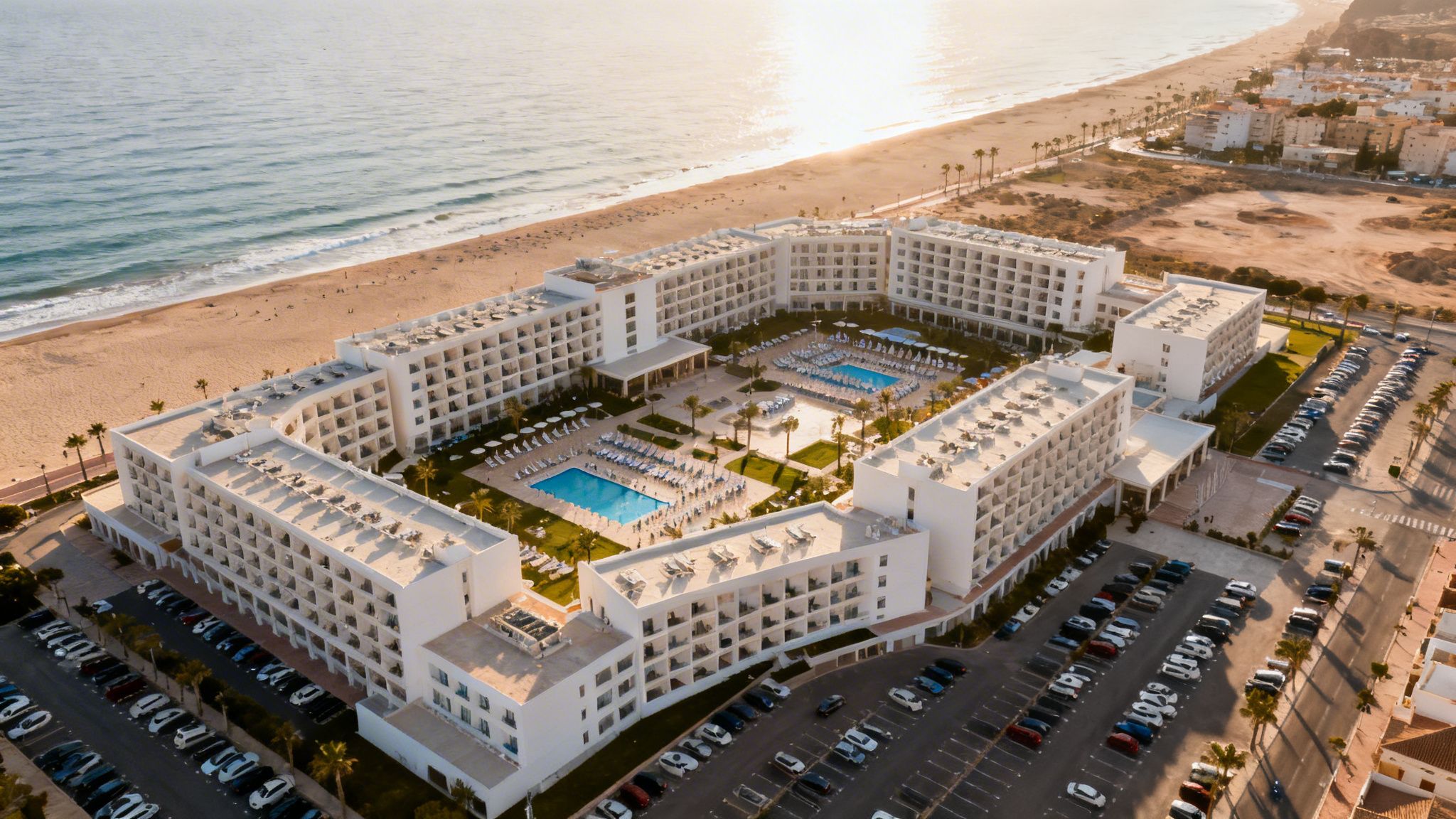 Vista aérea de un hotel blanco junto a una playa de arena con piscinas y palmeras al atardecer.
