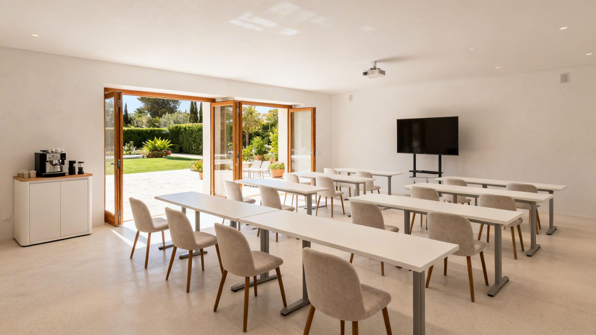 Sala de reuniones luminosa con mesas, sillas, pantalla, proyector y zona de café, con vistas a un jardín soleado.