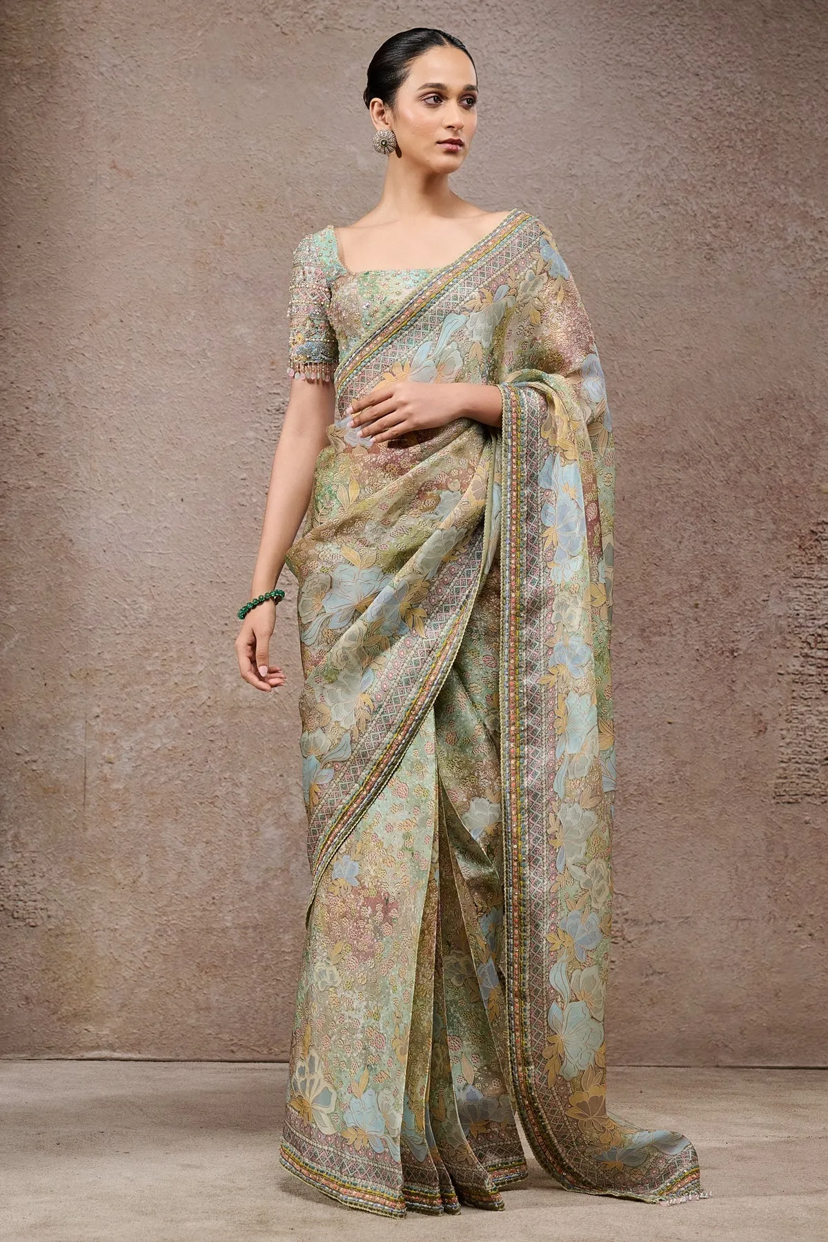 This pastel-toned sari is a vision of grace, featuring soft floral prints in shades of mint, blush, and sky blue. The border is intricately detailed with geometric motifs, lending a refined touch. Paired with an embellished blouse adorned with sequins and delicate beadwork, the ensemble beautifully blends traditional artistry with modern sophistication.