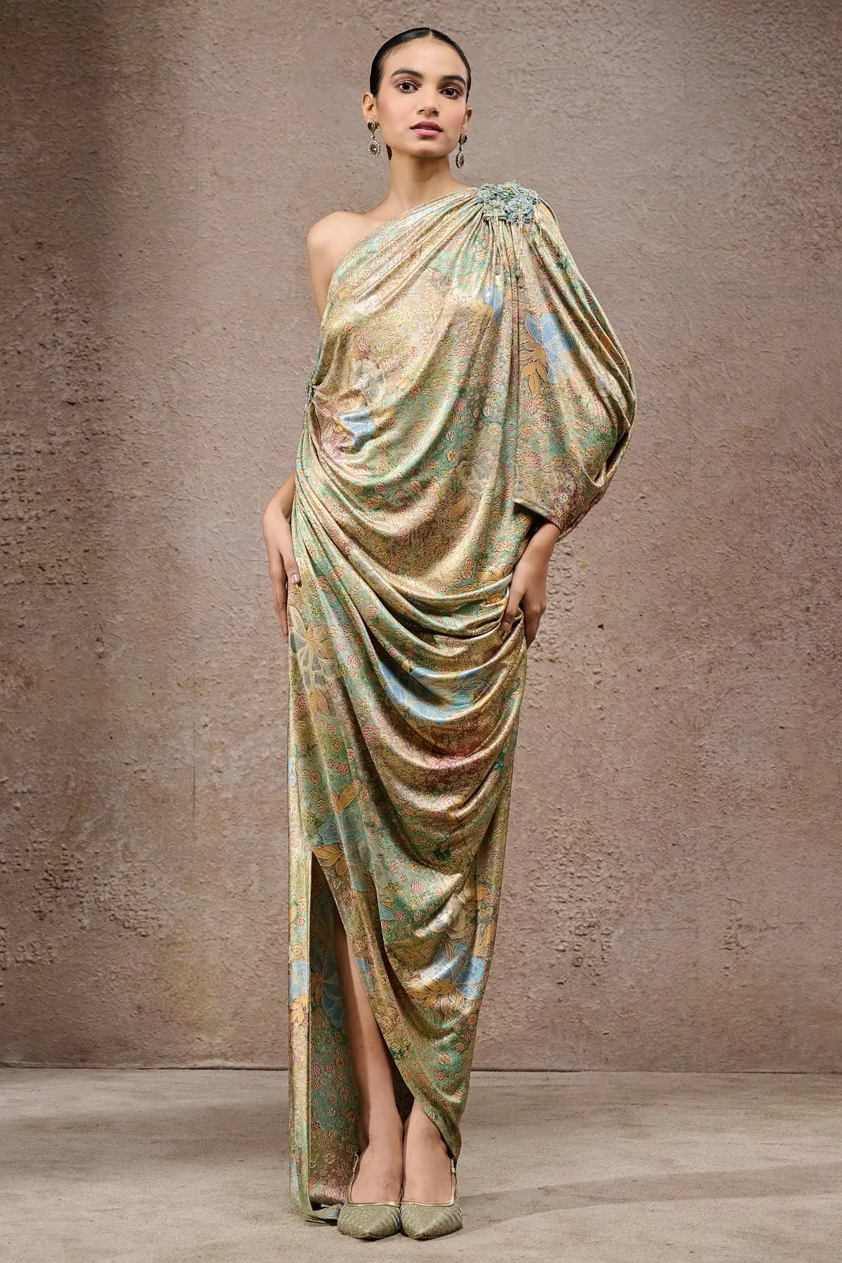 Effortlessly glamorous, this one-shoulder metallic draped gown showcases fluid drapery in lustrous fabric with pastel floral undertones. The asymmetrical silhouette is accentuated with gathered detailing at the shoulder, highlighted by an embellished floral brooch. A thigh-high slit adds a modern edge, making this gown a statement piece that fuses elegance with contemporary flair.