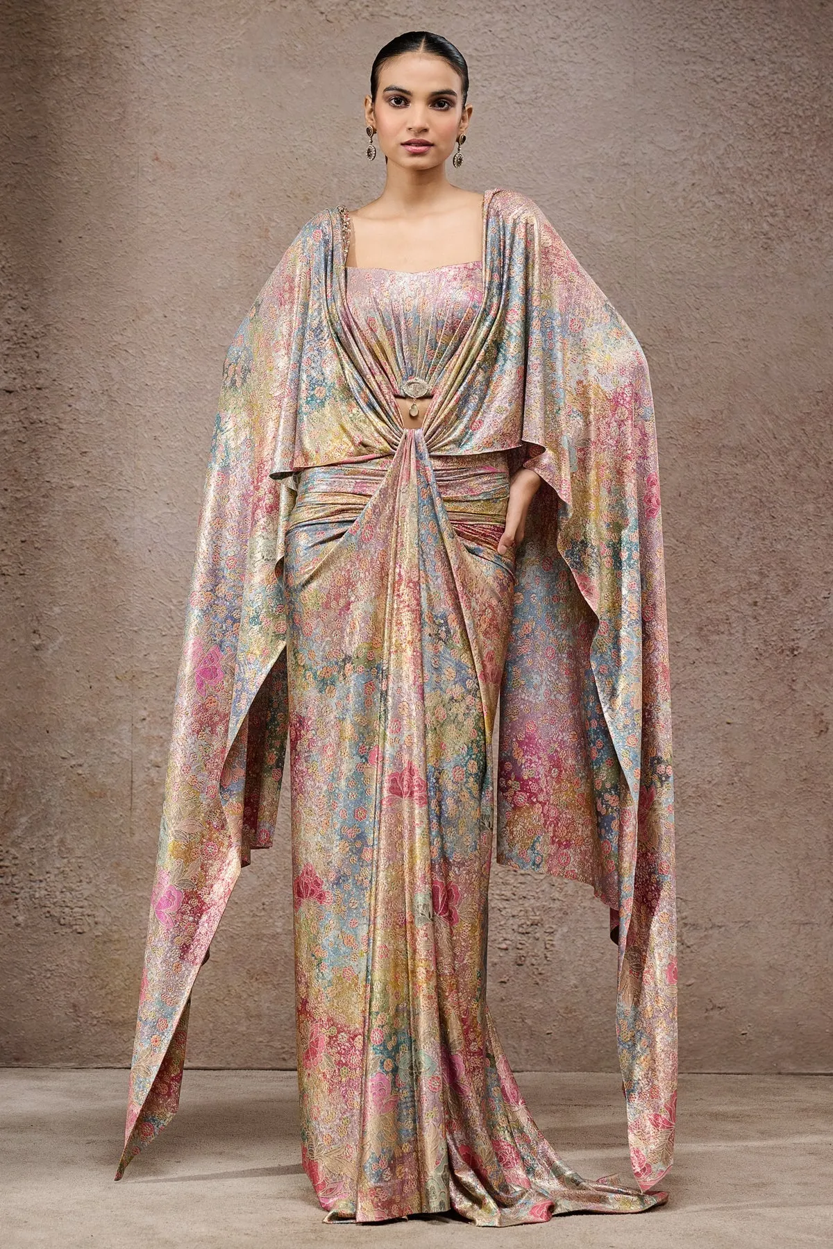 A contemporary take on the timeless sari, this metallic multicolor drape is crafted in fluid fabric with a lustrous finish. Adorned with abstract floral patterns, the design features structured draping with a gathered knot detail and statement brooch at the waist, enhancing its modern appeal. Flowing cape-style sleeves elevate the look with dramatic flair, making it a standout piece for glamorous occasions.