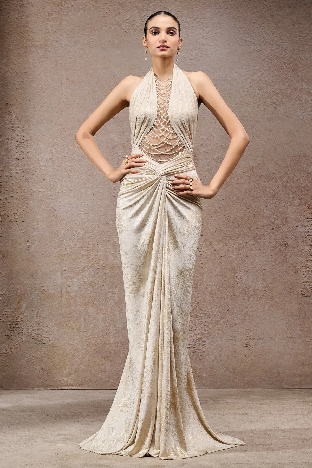 Effortlessly glamorous, this champagne gown is designed with a halter neckline and delicate pearl bead embellishments cascading over the sheer bodice. The silhouette features intricate draping at the waist, creating a figure-enhancing effect that flows into a floor-length skirt. Perfectly blending sophistication with allure, this gown makes a striking statement for evening soirées and celebratory parties.