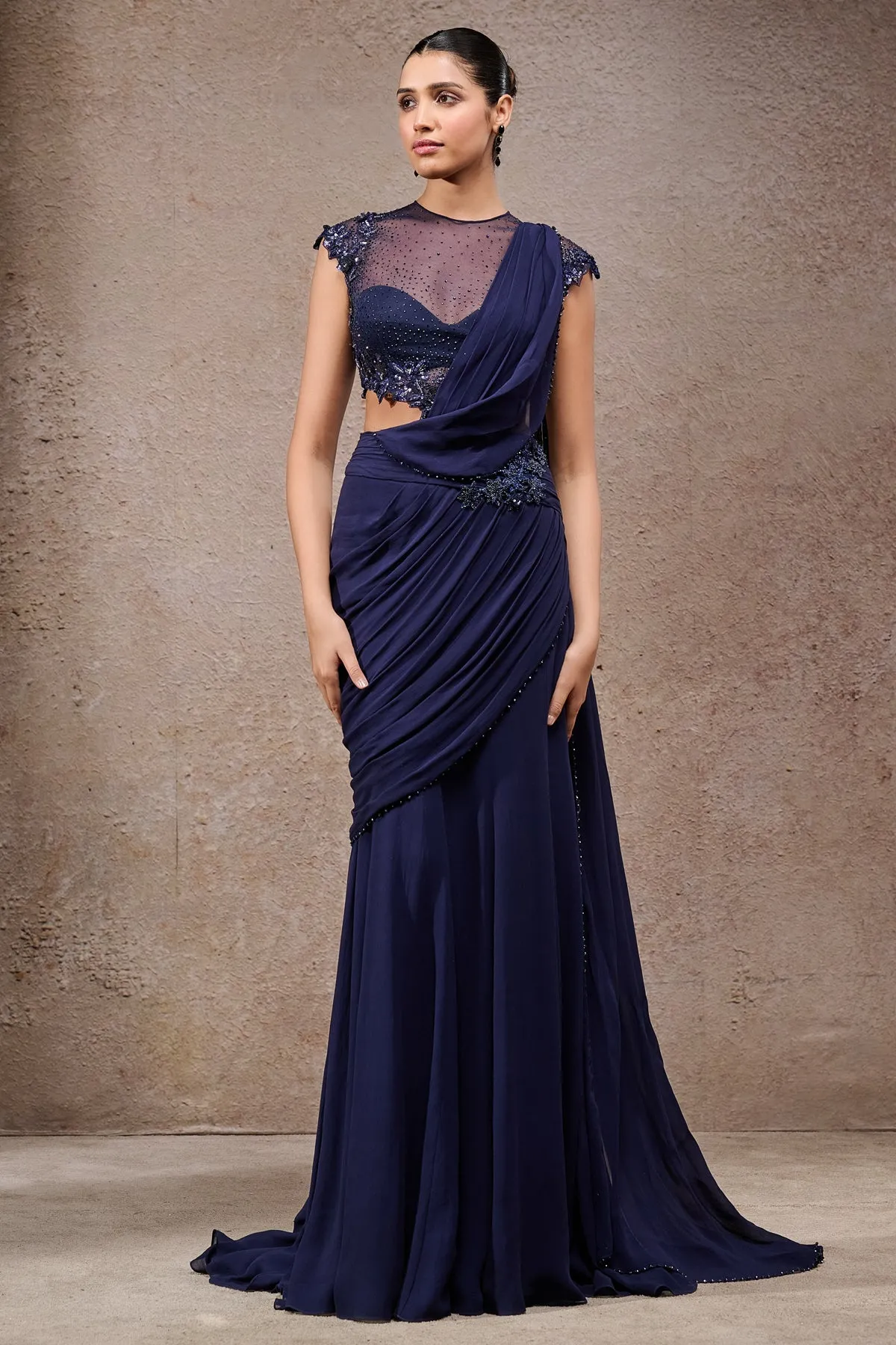 This contemporary navy blue sari exudes refined sophistication with its pre-draped silhouette. Featuring a sheer embellished blouse with intricate beadwork and cap sleeves, it blends traditional charm with modern elegance. The fluid drape cascades gracefully, creating a statement-making look perfect for festive and evening occasions.