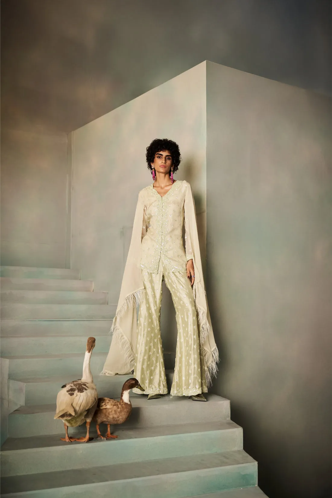 A vision of understated grace, this ivory sharara set is elevated with fine sequin accents that shimmer delicately against its soft fabric. The longline tunic is paired with flared sharara trousers, exuding timeless charm. A flowing draped cape with fringed edges cascades elegantly, completing the ensemble with a couture edge, perfect for soirées and celebrations.
