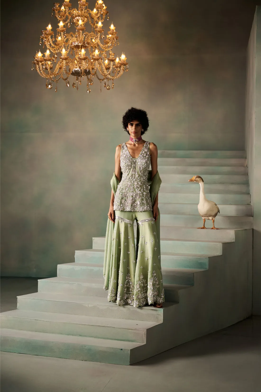 This sage green sharara set captures ethereal beauty with its shimmering silver embroidery and delicate sequin embellishments. The sleeveless, deep-neck tunic is intricately adorned with floral and vine motifs, perfectly paired with flowing sharara trousers featuring ornate borders. Draped with a soft matching dupatta, the ensemble radiates timeless grace and couture finesse, ideal for weddings and regal soirées.