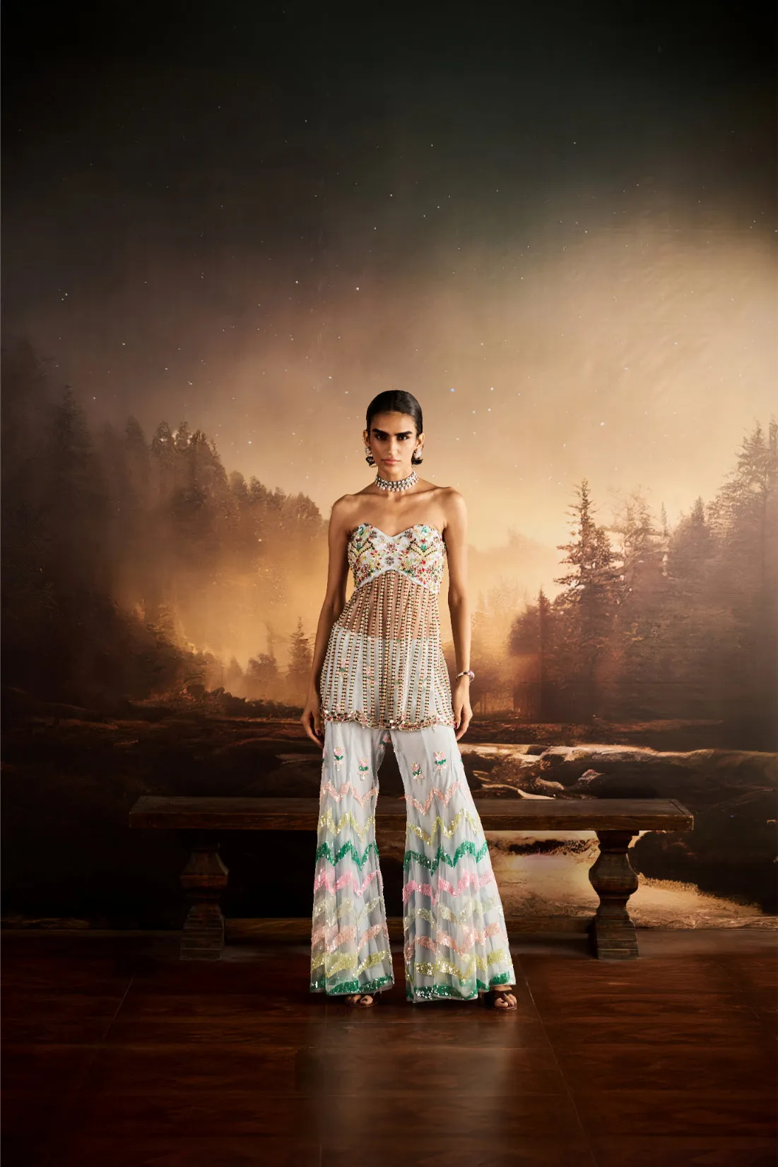 A playful yet refined creation, this sharara set brings together the vibrance of pastel hues with the elegance of couture craftsmanship. The strapless bustier is delicately adorned with floral embroidery, while the sheer overlay cascades with intricate detailing. Paired with flared sharara trousers embellished in chevron sequins and pastel embroidery, this ensemble is a modern celebration of color and artistry.