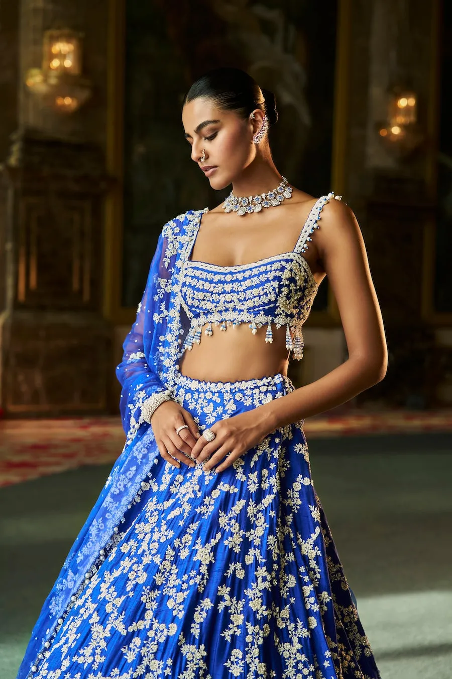 This emerald green lehenga is a luminous ode to nature-inspired couture, adorned with intricate ivory floral embroidery that cascades gracefully across the silhouette. The exquisitely crafted blouse features ornate handwork and delicate edging, creating a harmonious balance of structure and softness. A matching dupatta, lightly embellished, frames the ensemble with refined elegance and fluid movement. Fresh yet opulent, this creation embodies timeless artistry with a celebratory, regal presence.