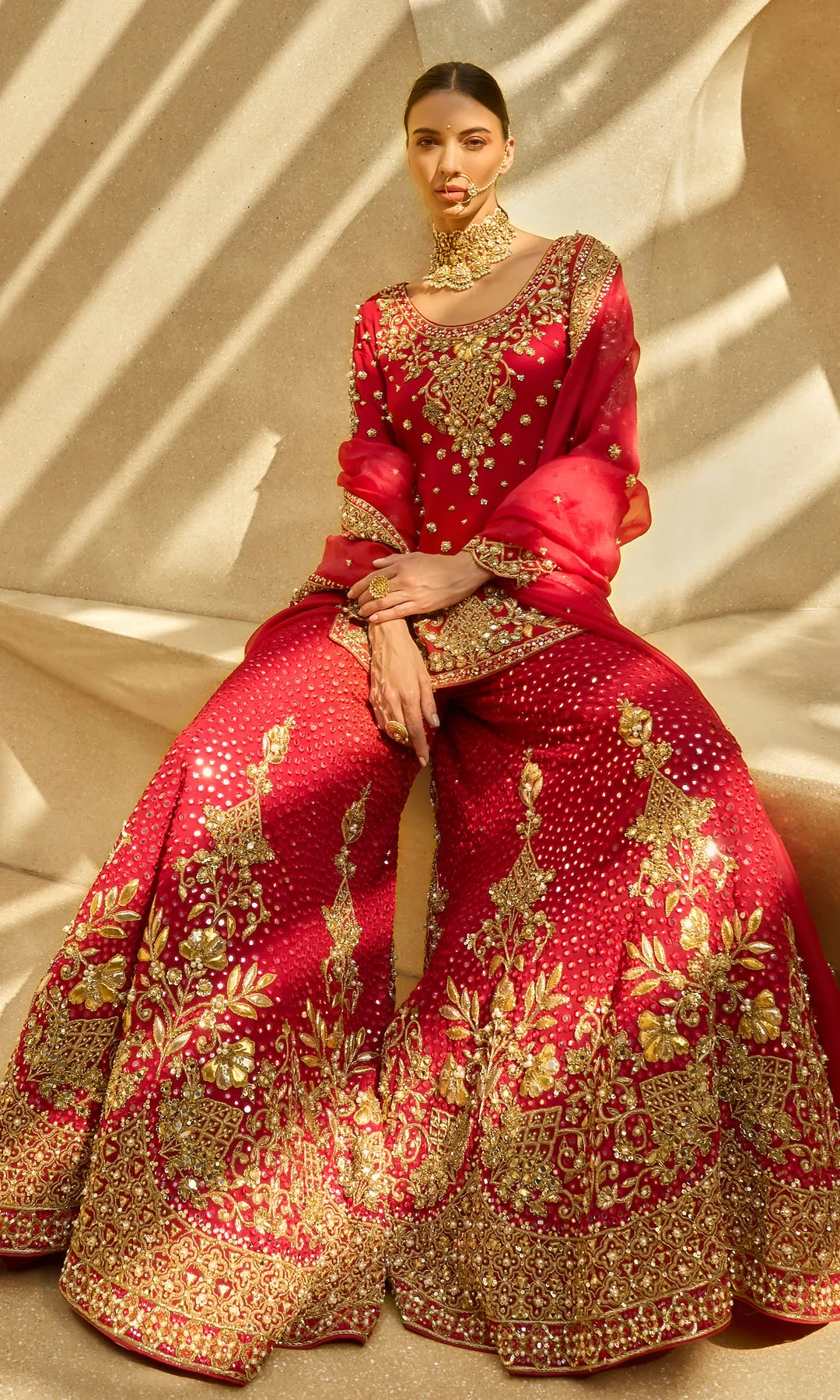 This opulent red lehenga is a timeless expression of bridal grandeur, crafted in a rich crimson hue and adorned with elaborate antique gold floral zardozi embroidery. The skirt features dense, heirloom-inspired motifs framed with a heavily embellished border, creating depth and ceremonial richness. The fitted blouse mirrors the intricate craftsmanship with ornate detailing and a structured silhouette, while the sheer red dupatta edged in gold adds regal movement and traditional elegance. Designed to embody classic bridal splendor with enduring sophistication, this ensemble is a celebration of heritage, artistry, and majestic presence.