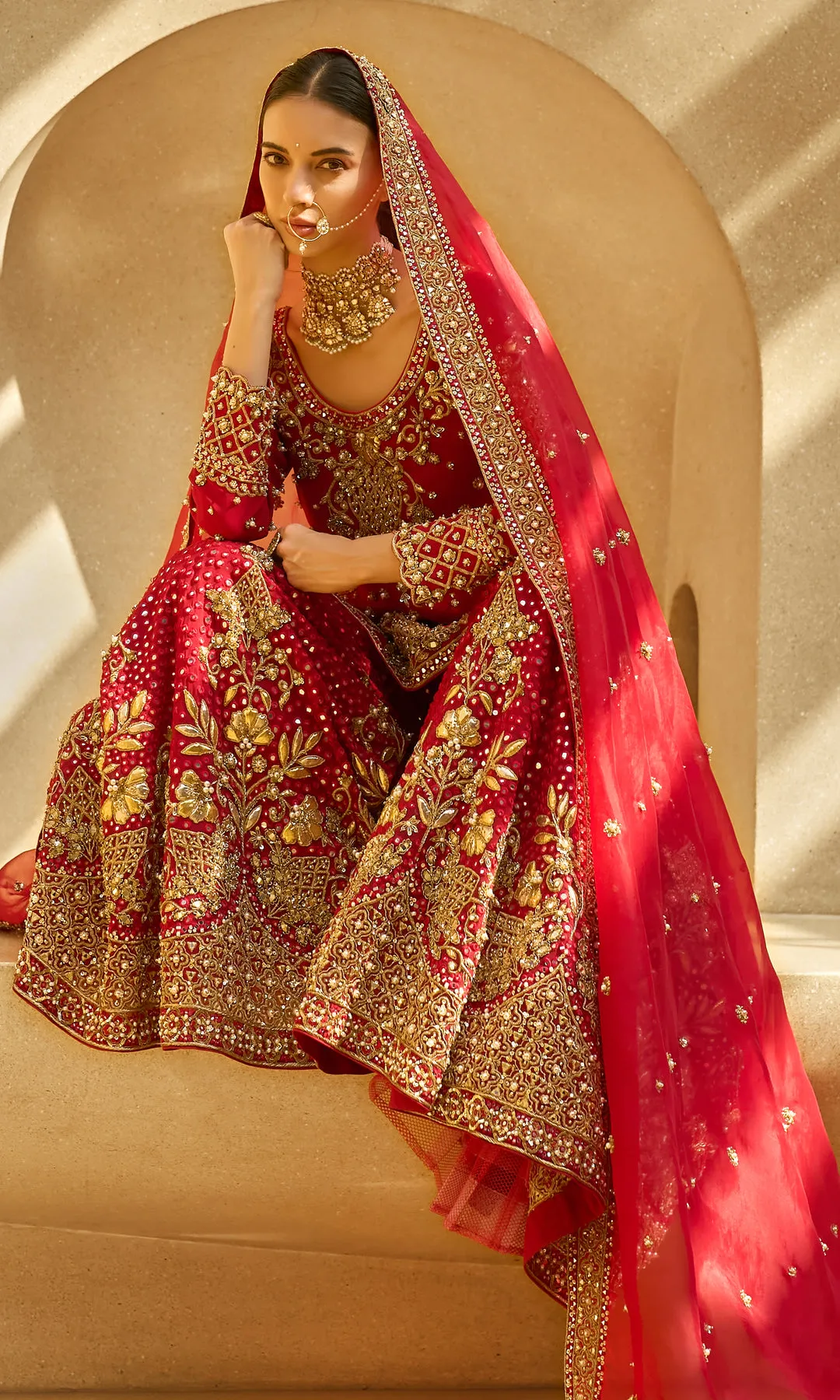 This opulent red lehenga is a timeless expression of bridal grandeur, crafted in a rich crimson hue and adorned with elaborate antique gold floral zardozi embroidery. The skirt features dense, heirloom-inspired motifs framed with a heavily embellished border, creating depth and ceremonial richness. The fitted blouse mirrors the intricate craftsmanship with ornate detailing and a structured silhouette, while the sheer red dupatta edged in gold adds regal movement and traditional elegance. Designed to embody classic bridal splendor with enduring sophistication, this ensemble is a celebration of heritage, artistry, and majestic presence.