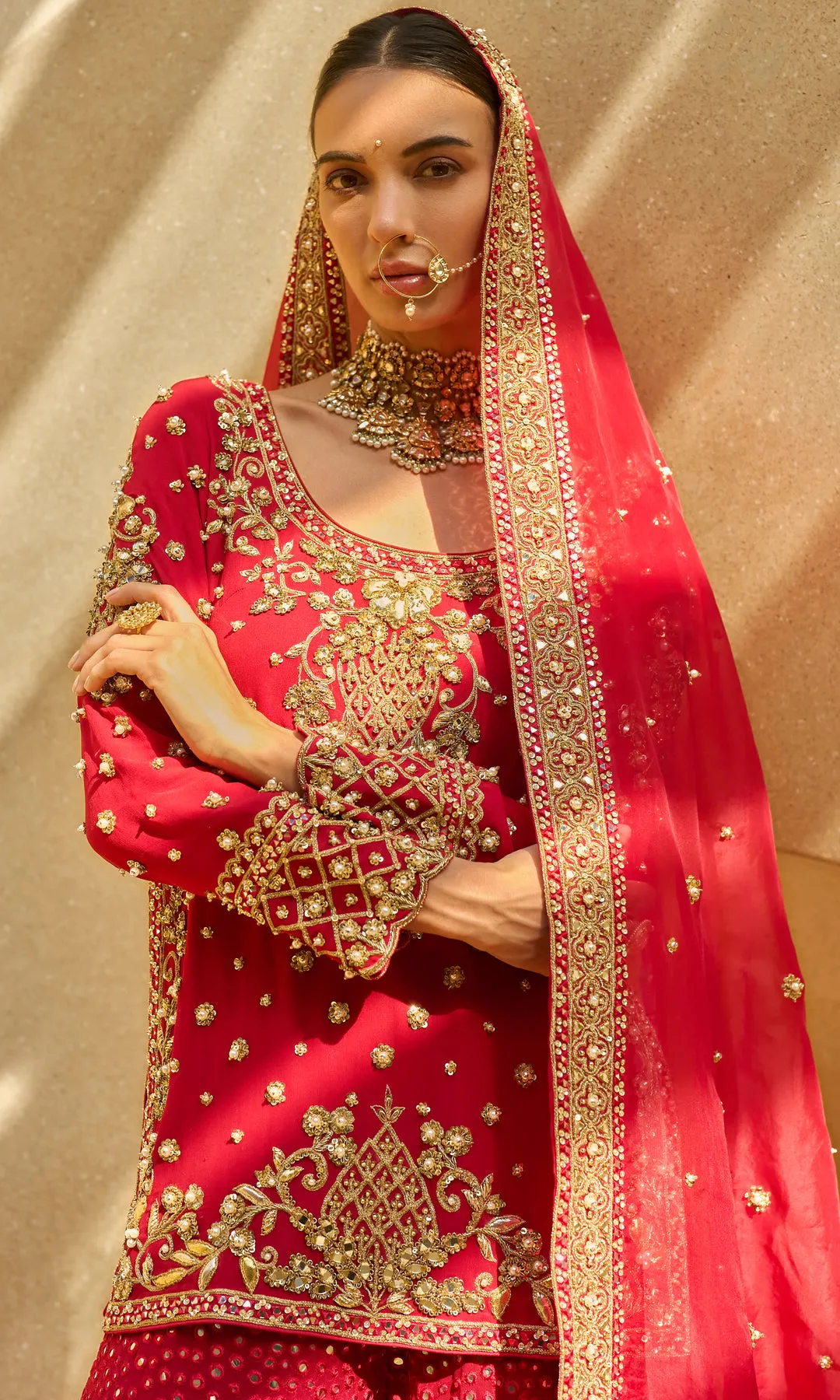 This opulent red lehenga is a timeless expression of bridal grandeur, crafted in a rich crimson hue and adorned with elaborate antique gold floral zardozi embroidery. The skirt features dense, heirloom-inspired motifs framed with a heavily embellished border, creating depth and ceremonial richness. The fitted blouse mirrors the intricate craftsmanship with ornate detailing and a structured silhouette, while the sheer red dupatta edged in gold adds regal movement and traditional elegance. Designed to embody classic bridal splendor with enduring sophistication, this ensemble is a celebration of heritage, artistry, and majestic presence.