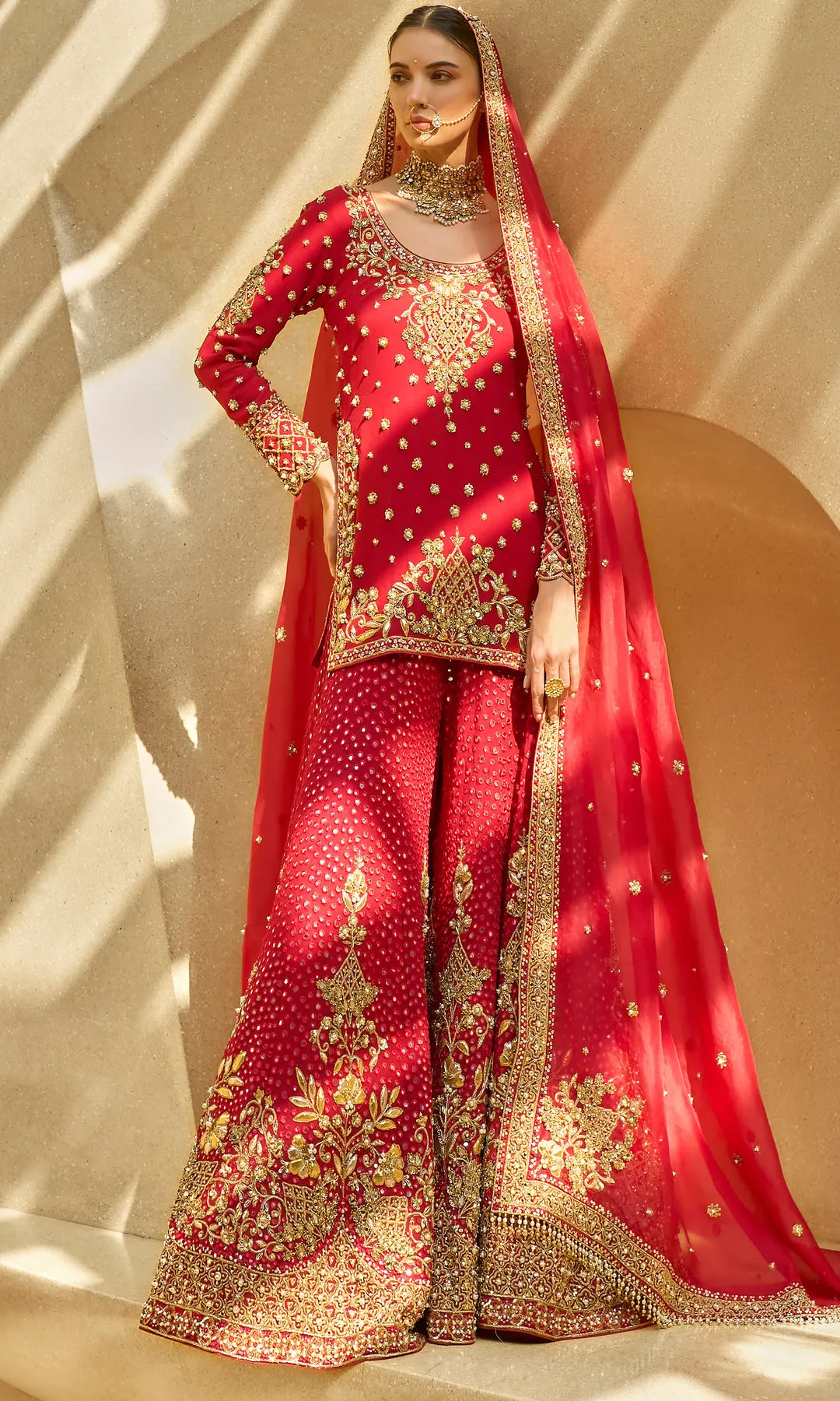 This opulent red lehenga is a timeless expression of bridal grandeur, crafted in a rich crimson hue and adorned with elaborate antique gold floral zardozi embroidery. The skirt features dense, heirloom-inspired motifs framed with a heavily embellished border, creating depth and ceremonial richness. The fitted blouse mirrors the intricate craftsmanship with ornate detailing and a structured silhouette, while the sheer red dupatta edged in gold adds regal movement and traditional elegance. Designed to embody classic bridal splendor with enduring sophistication, this ensemble is a celebration of heritage, artistry, and majestic presence.