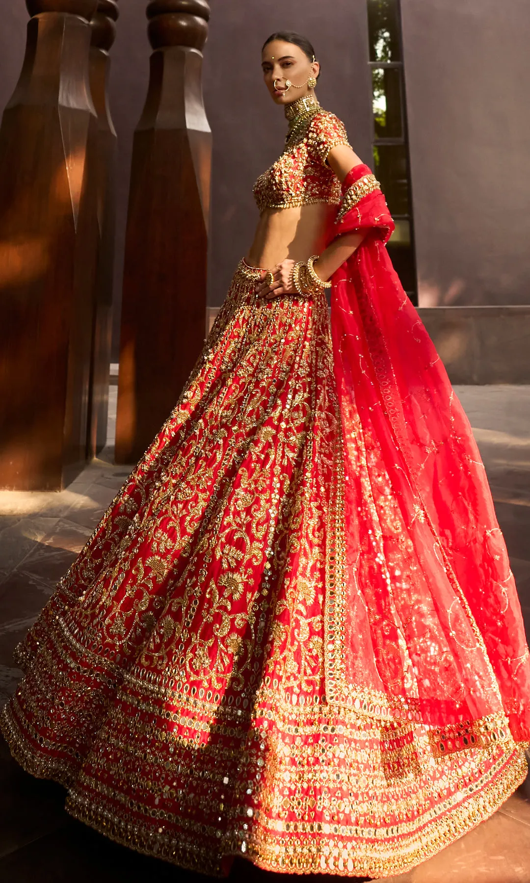 This striking deep crimson lehenga is a celebration of precision craftsmanship and royal elegance. The skirt features dense, all-over chevron and paisley zardozi embroidery in antique gold, creating a rhythmic geometric flow that enhances its voluminous silhouette. The structured blouse is richly embellished to match, with a sculpted neckline that adds modern strength to a classic form. Accented with heritage jewelry styling and a seamless finish, this ensemble commands attention through its bold color, architectural embroidery, and ceremonial grandeur—crafted for a bride who embodies power, grace, and tradition.