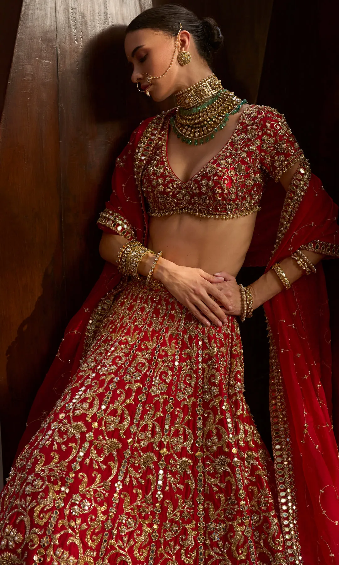 This striking deep crimson lehenga is a celebration of precision craftsmanship and royal elegance. The skirt features dense, all-over chevron and paisley zardozi embroidery in antique gold, creating a rhythmic geometric flow that enhances its voluminous silhouette. The structured blouse is richly embellished to match, with a sculpted neckline that adds modern strength to a classic form. Accented with heritage jewelry styling and a seamless finish, this ensemble commands attention through its bold color, architectural embroidery, and ceremonial grandeur—crafted for a bride who embodies power, grace, and tradition.