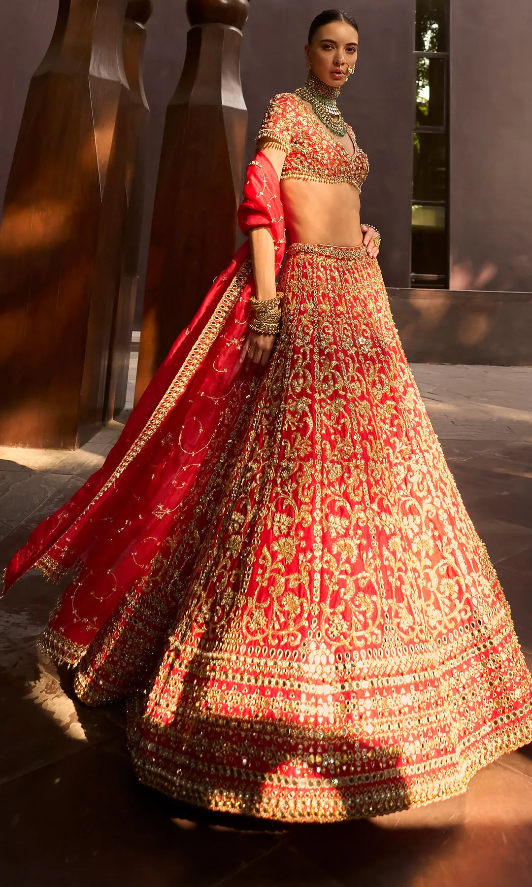 This striking deep crimson lehenga is a celebration of precision craftsmanship and royal elegance. The skirt features dense, all-over chevron and paisley zardozi embroidery in antique gold, creating a rhythmic geometric flow that enhances its voluminous silhouette. The structured blouse is richly embellished to match, with a sculpted neckline that adds modern strength to a classic form. Accented with heritage jewelry styling and a seamless finish, this ensemble commands attention through its bold color, architectural embroidery, and ceremonial grandeur—crafted for a bride who embodies power, grace, and tradition.