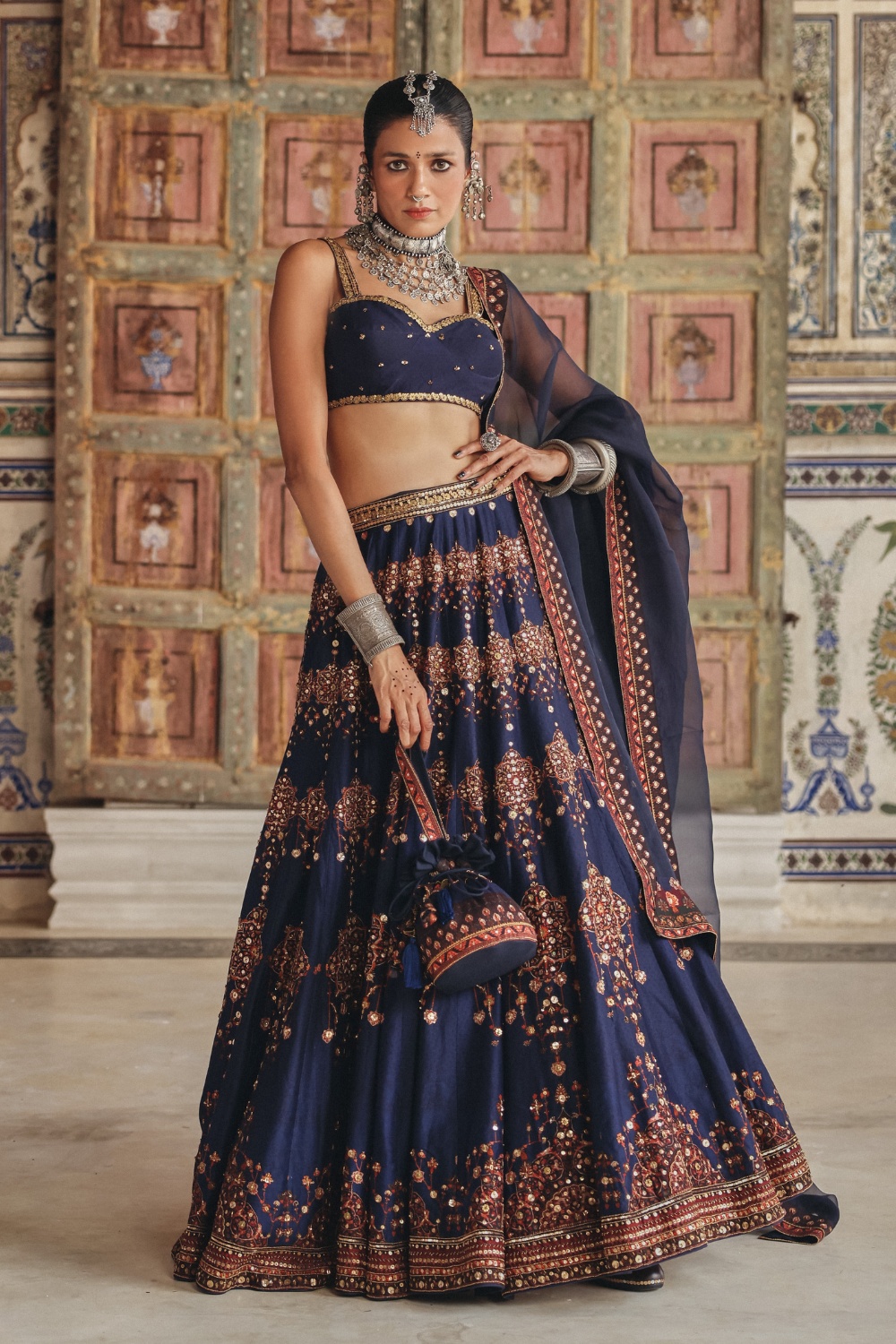 Exuding regal charm, this midnight navy sharara set is beautifully adorned with delicate gold-toned motifs and intricate embroidery that celebrate timeless craftsmanship. The kurta features an ornate embroidered neckline enriched with traditional zardozi-inspired detailing, while the flowing sharara is accentuated with heavily embellished borders showcasing mirror-inspired accents and fine threadwork. Paired with a coordinating dupatta framed by heritage-inspired geometric borders and subtle embellishments, the ensemble embodies understated luxury. The rich navy hue contrasted with antique gold detailing makes it an elegant choice for festive occasions and evening celebrations.