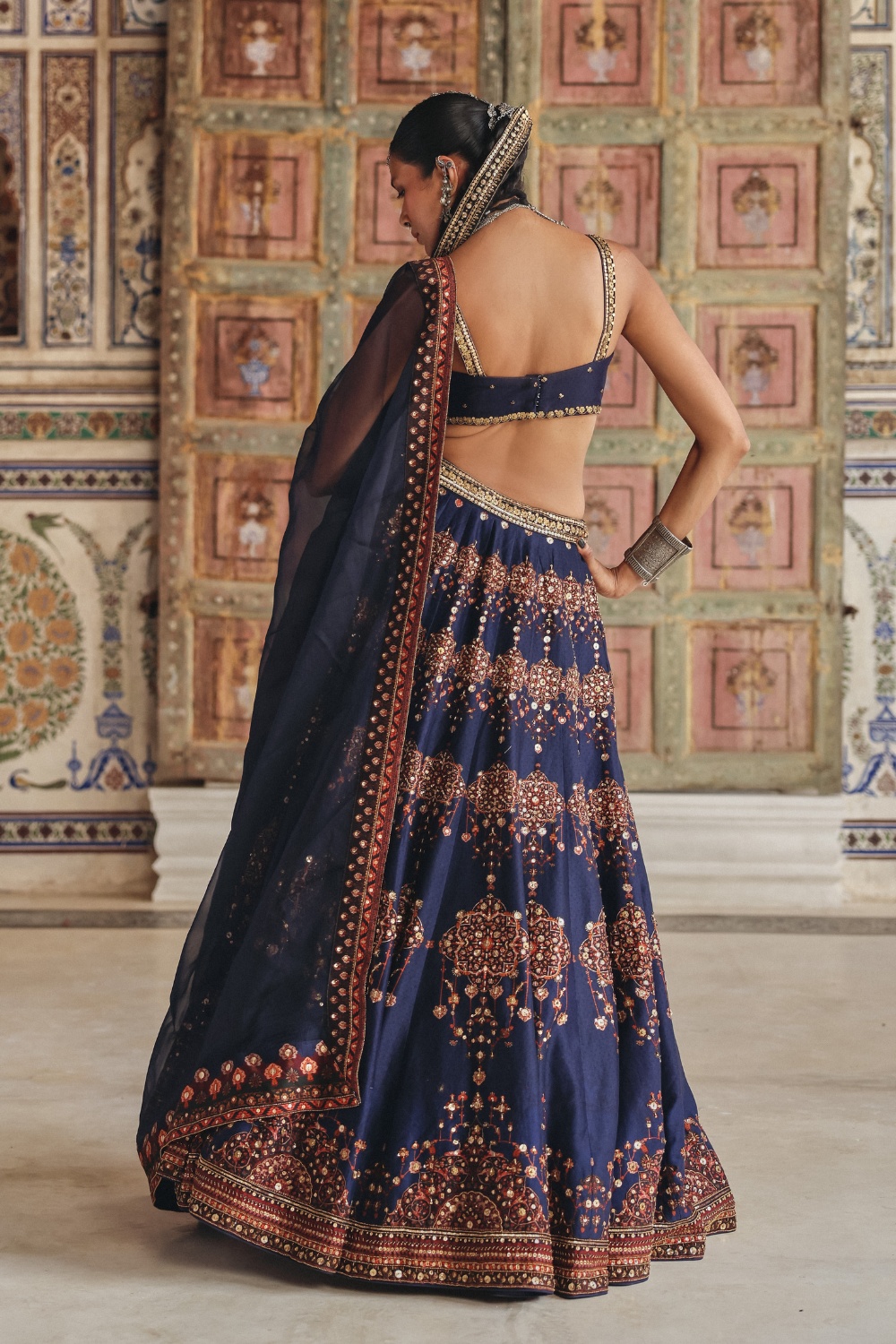 Exuding regal charm, this midnight navy sharara set is beautifully adorned with delicate gold-toned motifs and intricate embroidery that celebrate timeless craftsmanship. The kurta features an ornate embroidered neckline enriched with traditional zardozi-inspired detailing, while the flowing sharara is accentuated with heavily embellished borders showcasing mirror-inspired accents and fine threadwork. Paired with a coordinating dupatta framed by heritage-inspired geometric borders and subtle embellishments, the ensemble embodies understated luxury. The rich navy hue contrasted with antique gold detailing makes it an elegant choice for festive occasions and evening celebrations.