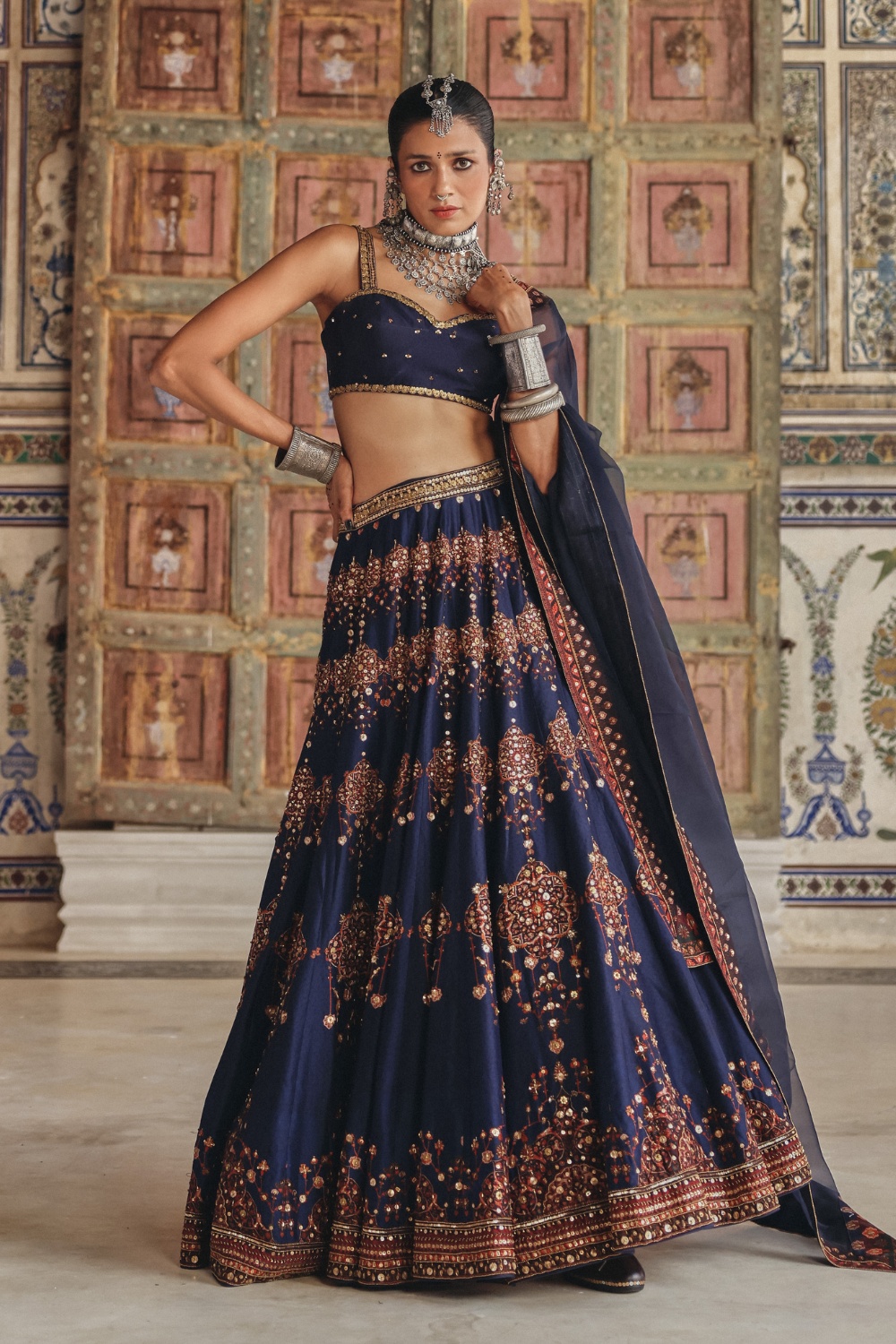 Exuding regal charm, this midnight navy sharara set is beautifully adorned with delicate gold-toned motifs and intricate embroidery that celebrate timeless craftsmanship. The kurta features an ornate embroidered neckline enriched with traditional zardozi-inspired detailing, while the flowing sharara is accentuated with heavily embellished borders showcasing mirror-inspired accents and fine threadwork. Paired with a coordinating dupatta framed by heritage-inspired geometric borders and subtle embellishments, the ensemble embodies understated luxury. The rich navy hue contrasted with antique gold detailing makes it an elegant choice for festive occasions and evening celebrations.