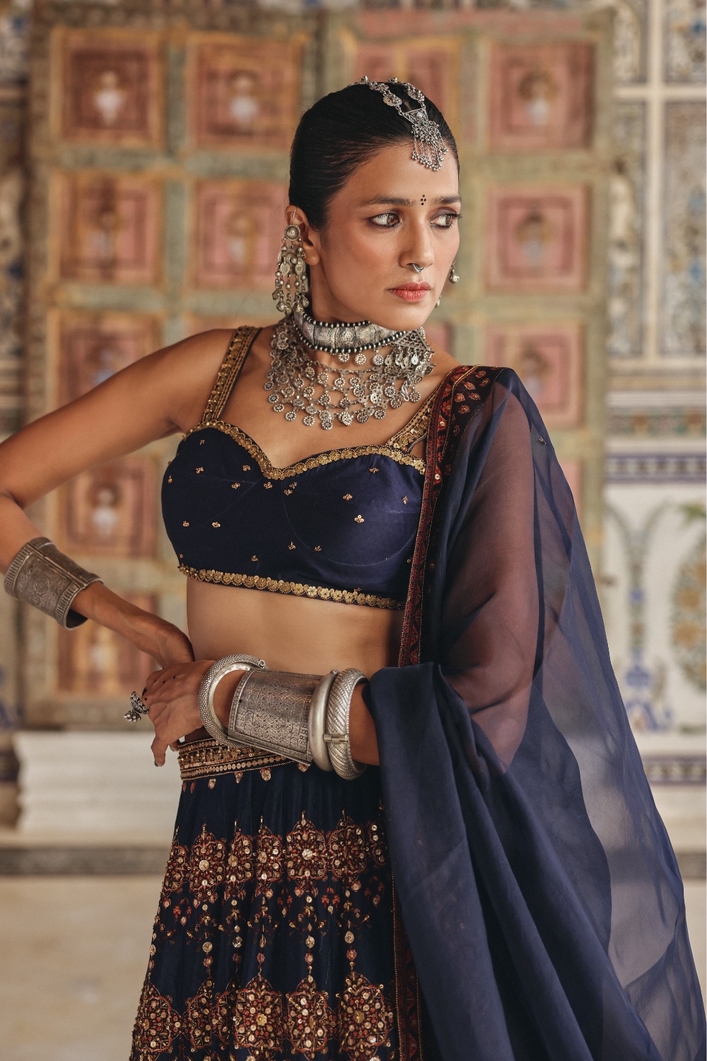 Exuding regal charm, this midnight navy sharara set is beautifully adorned with delicate gold-toned motifs and intricate embroidery that celebrate timeless craftsmanship. The kurta features an ornate embroidered neckline enriched with traditional zardozi-inspired detailing, while the flowing sharara is accentuated with heavily embellished borders showcasing mirror-inspired accents and fine threadwork. Paired with a coordinating dupatta framed by heritage-inspired geometric borders and subtle embellishments, the ensemble embodies understated luxury. The rich navy hue contrasted with antique gold detailing makes it an elegant choice for festive occasions and evening celebrations.