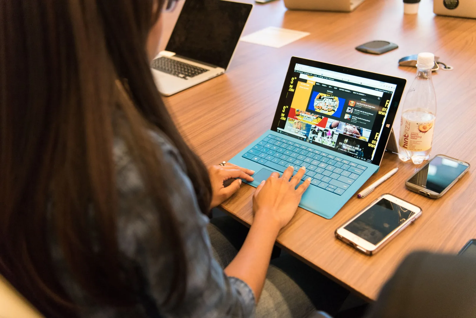 Person arbeitet an einem Tablet mit Tastatur an einem Besprechungstisch, daneben Smartphones und Laptop