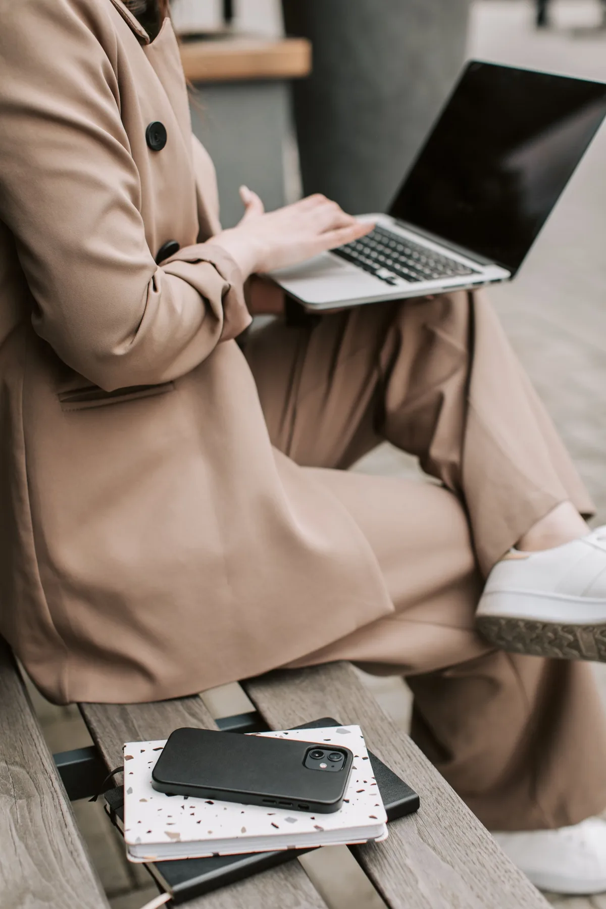 Person working on a laptop on a bench, with a smartphone and notebook beside them