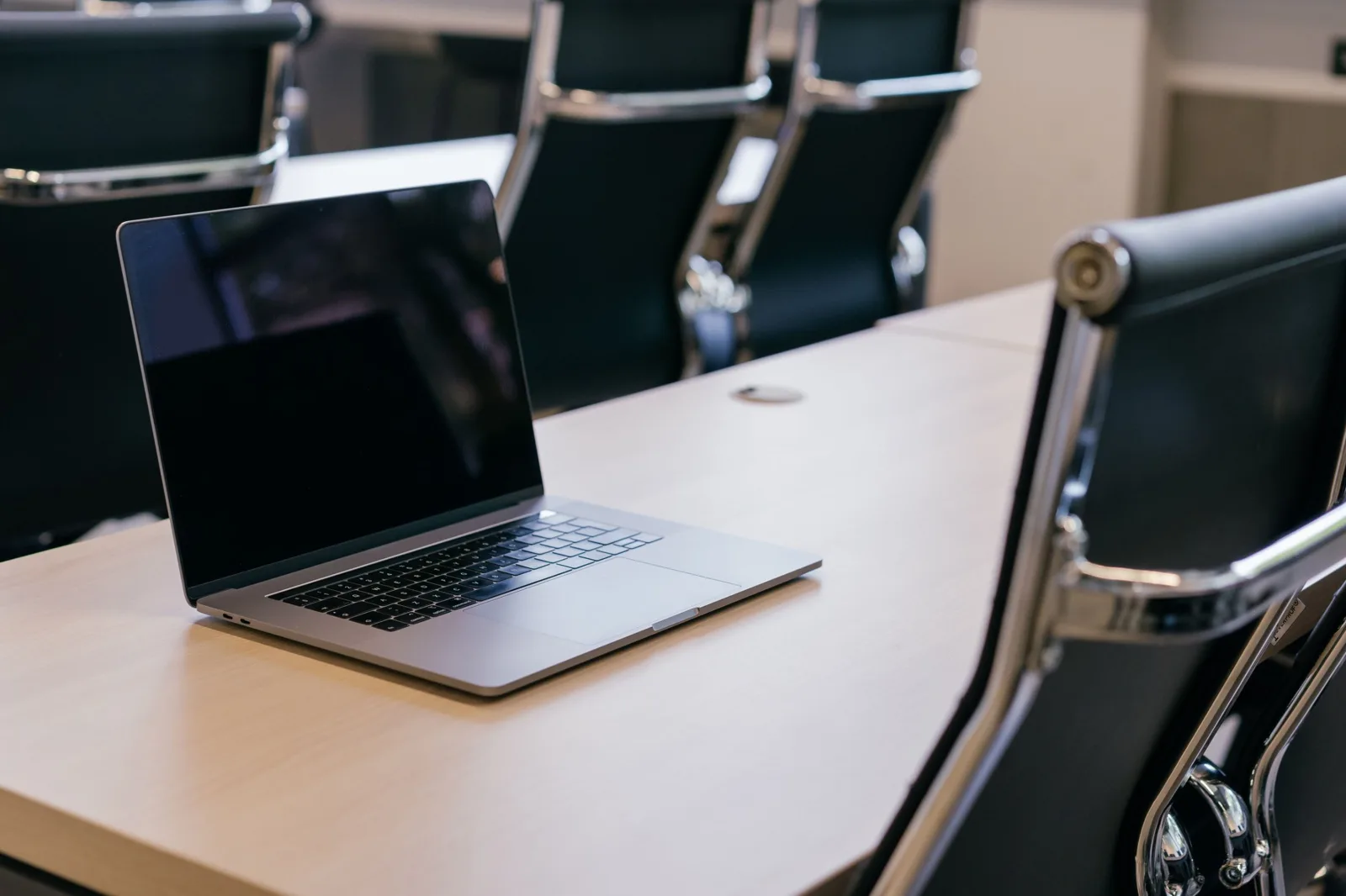 Open laptop on a light wooden table between black leather chairs in a meeting room
