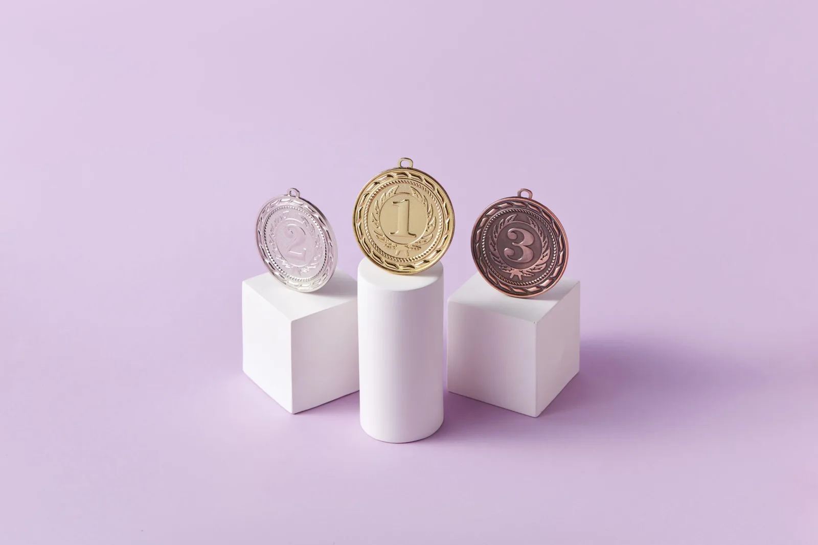 Gold-, Silber- und Bronzemedaille auf weißen Podesten vor einem fliederfarbenen Hintergrund