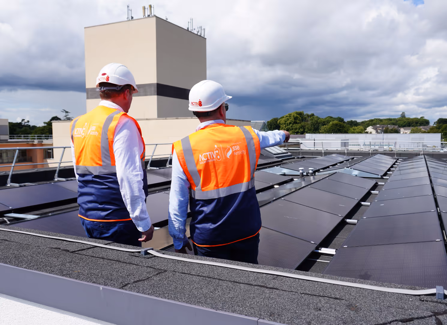 Two Activ8 SSE Renewables workers in safety gear inspect rooftop solar panel installation under a cloudy sky.