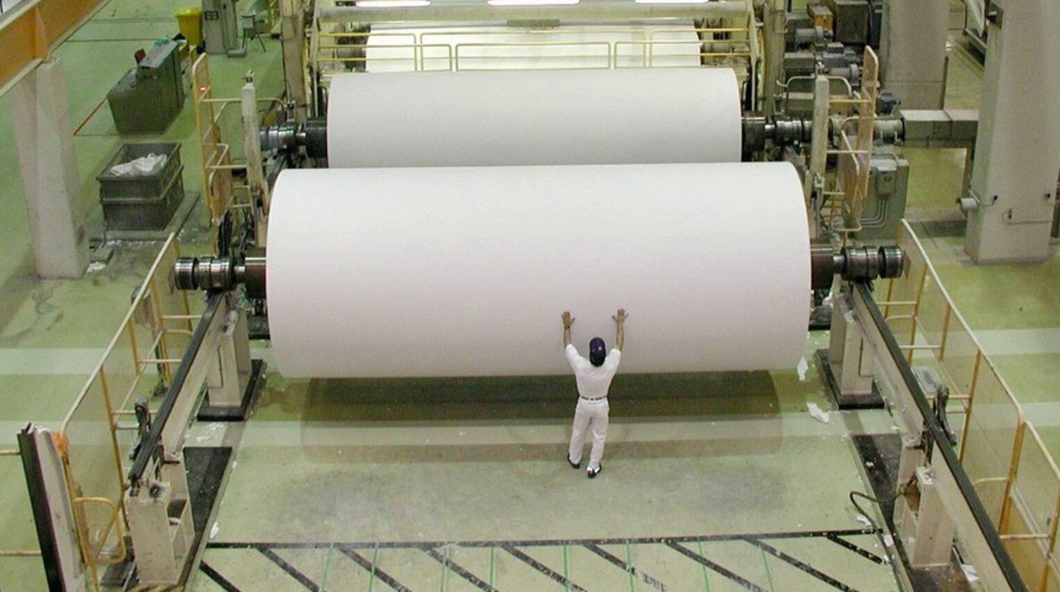 A worker dwarfed by giant paper rolls inside a paper mill