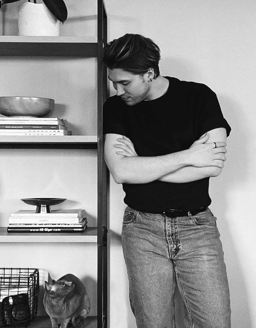 Liam in his home, smiling down at his cat that is sitting on a bookshelf below him.