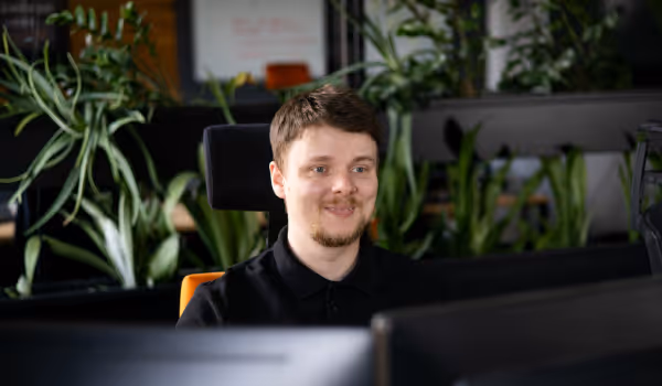 Mężczyzna siedzący przy biurku w nowoczesnym biurze, przed dwoma monitorami, z roślinami w tle.