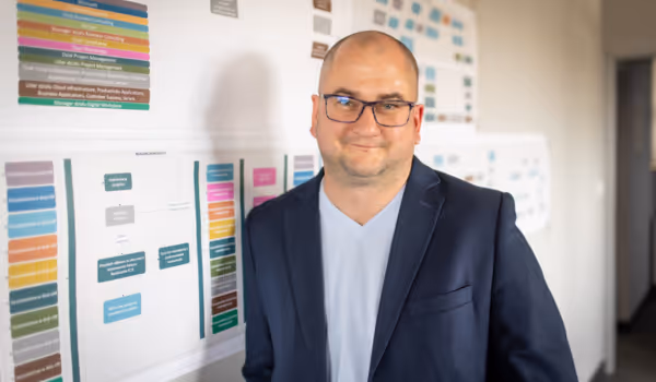 Mężczyzna w okularach i granatowej marynarce stoi przy ścianie z wykresami i diagramami w biurze.