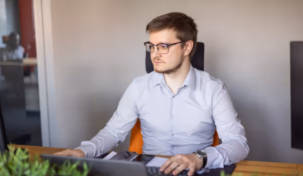 Mężczyzna w okularach i jasnej koszuli pracuje przy komputerze przy drewnianym biurku w jasnym biurze.